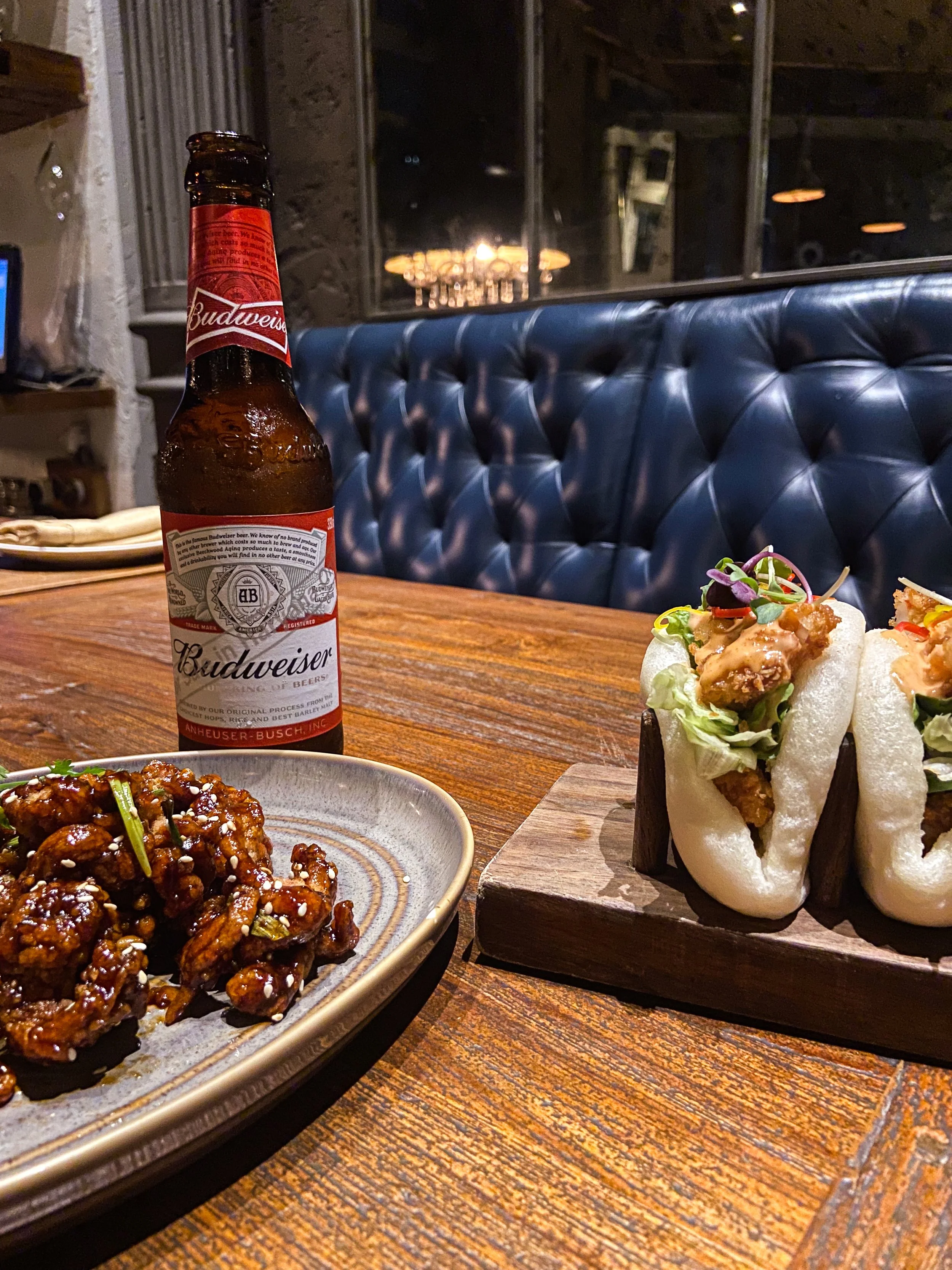 A bottle of Budweiser beer, a plate of glazed chicken wings with sesame seeds, and two steamed bao buns filled with fried chicken, lettuce, and sauce on a wooden table in a restaurant.