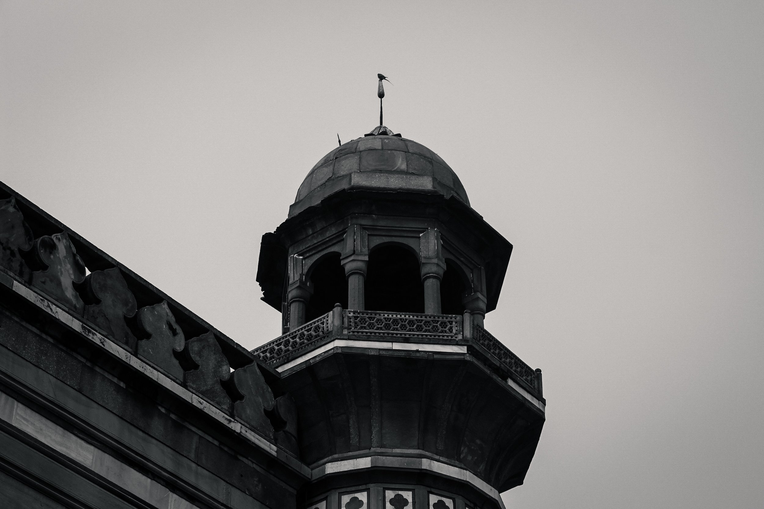 Black and white photo of a historic building's ornate tower with a decorative balcony and a weather vane in the shape of a bird on top, against a cloudy sky.