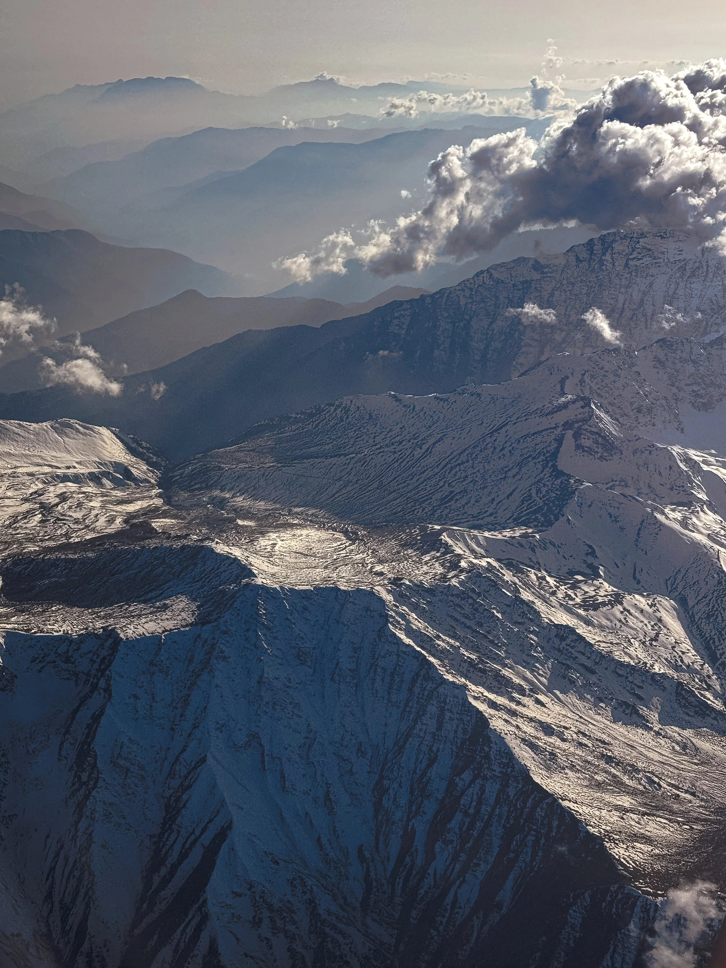 Aerial view of snow-capped mountains with multiple mountain ranges fading into the distance under a cloudy sky.