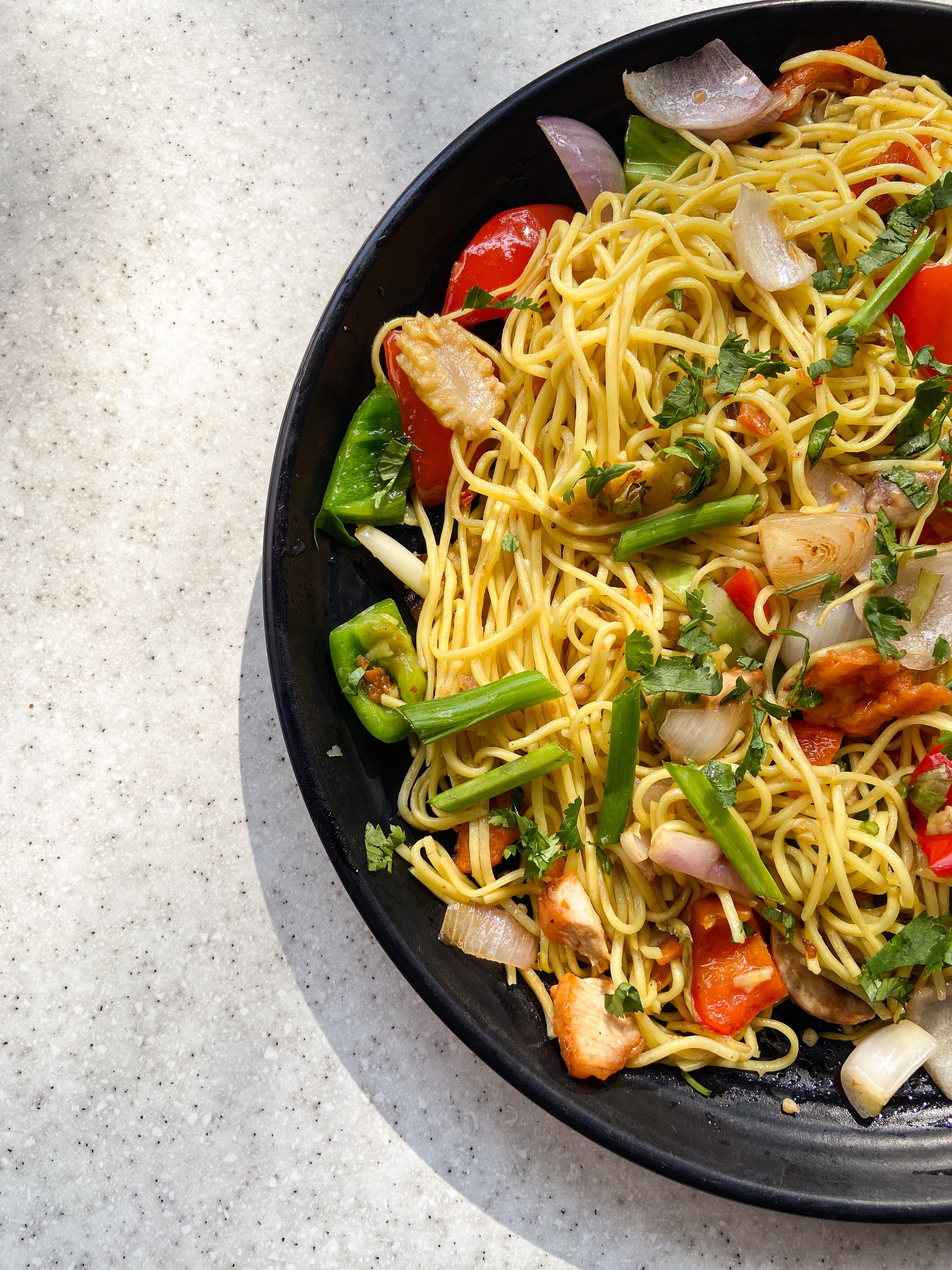 A plate of cooked ramen noodles with vegetables and herbs on a speckled light-colored surface.