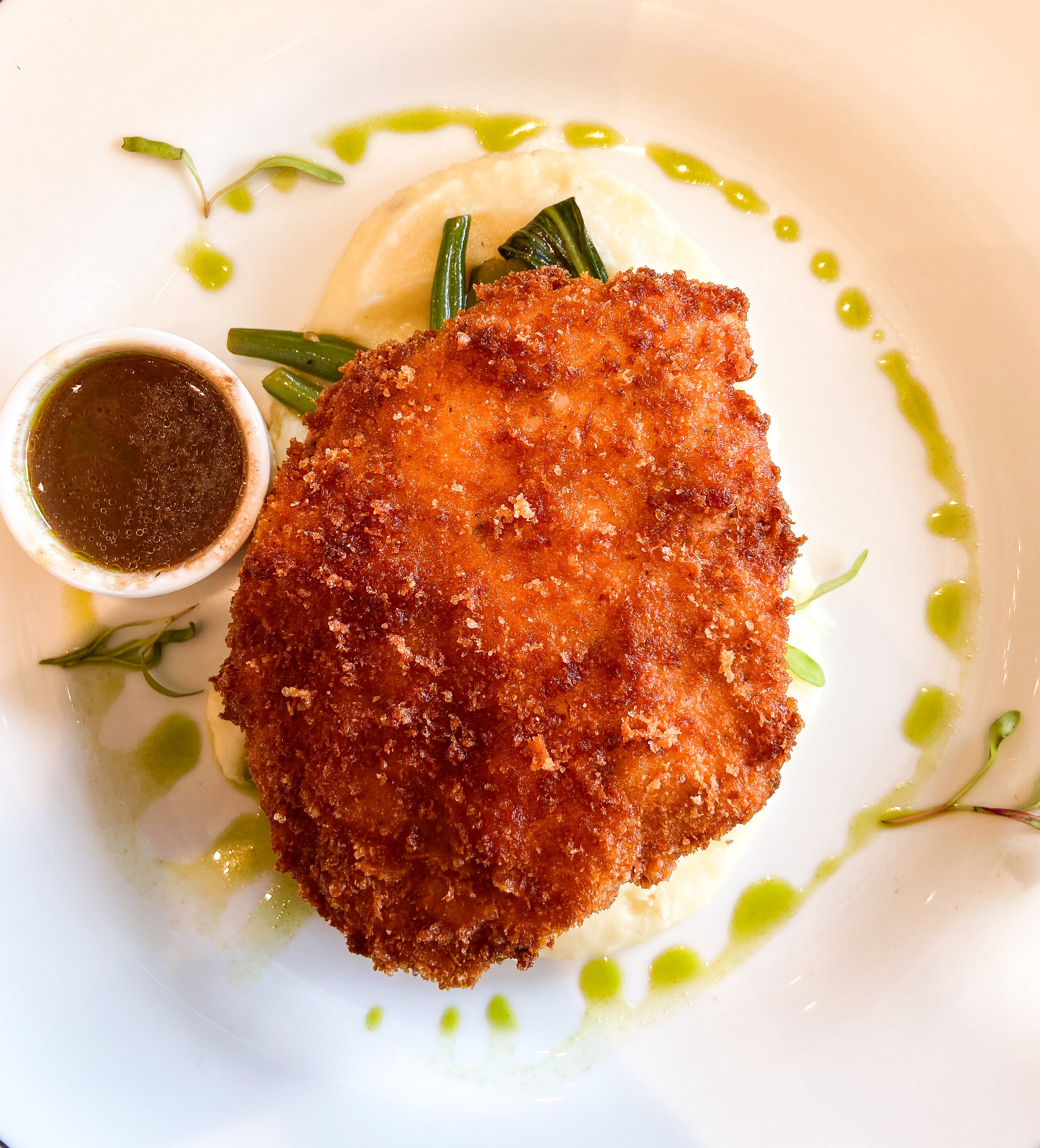 Fried fish filet with green beans, mashed potatoes, a small cup of gravy, and green sauce drizzled on the plate.