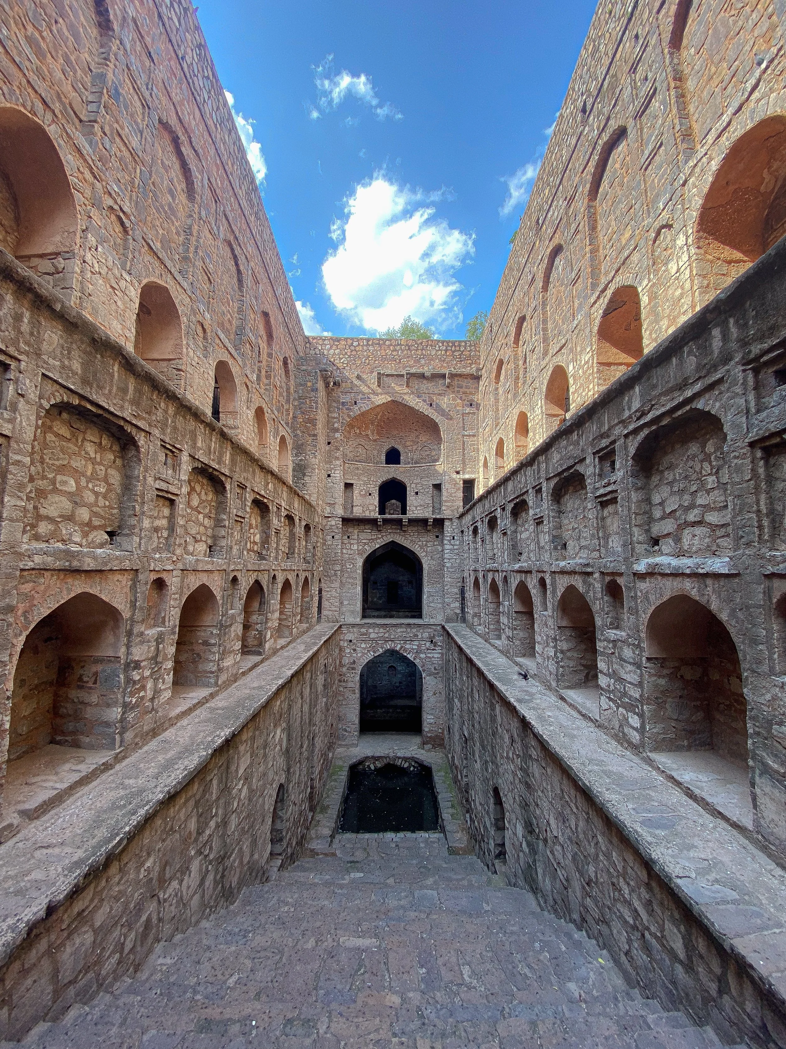 An ancient stone fortress with multiple arched windows and doorways, featuring a central water tank or moat at the bottom and a blue sky with clouds overhead.
