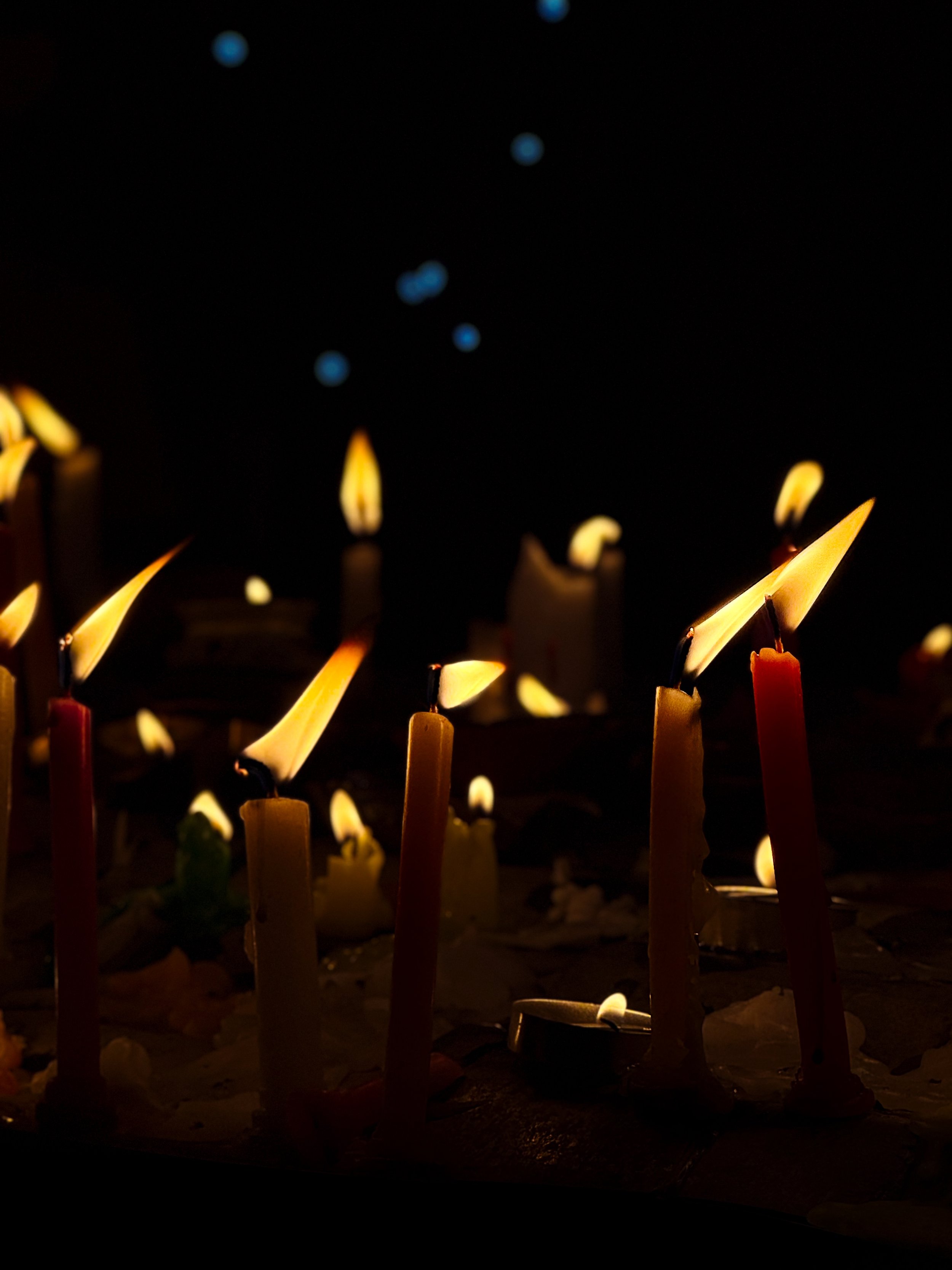 Several lit candles with melted wax on a dark surface, some with tiny flames, and a dark background with faint blue light spots.