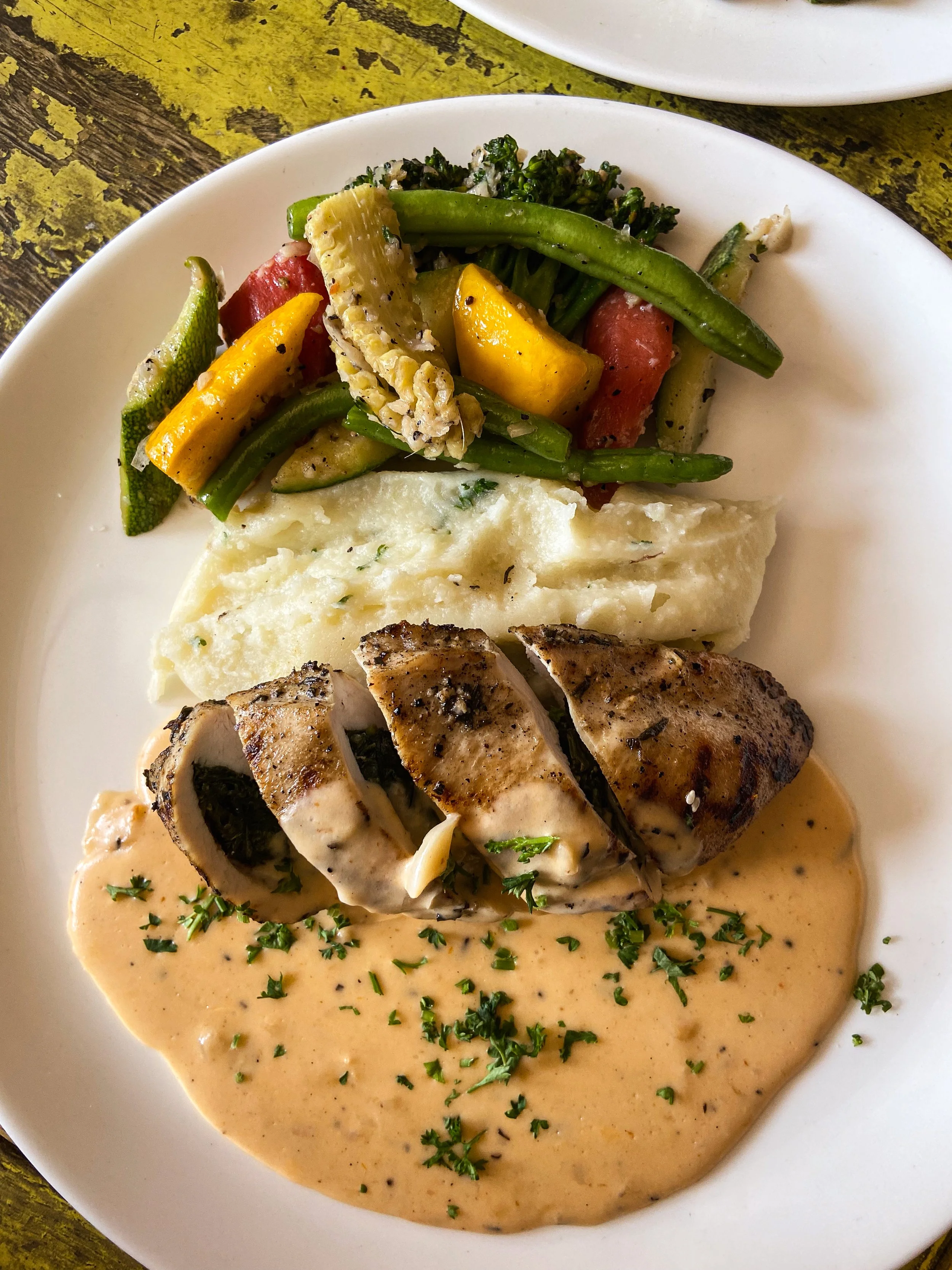 Plate with grilled fish fillet topped with creamy sauce, mashed potatoes, grilled vegetables, and a side of mixed vegetables including broccoli, green beans, zucchini, yellow squash, and tomato.