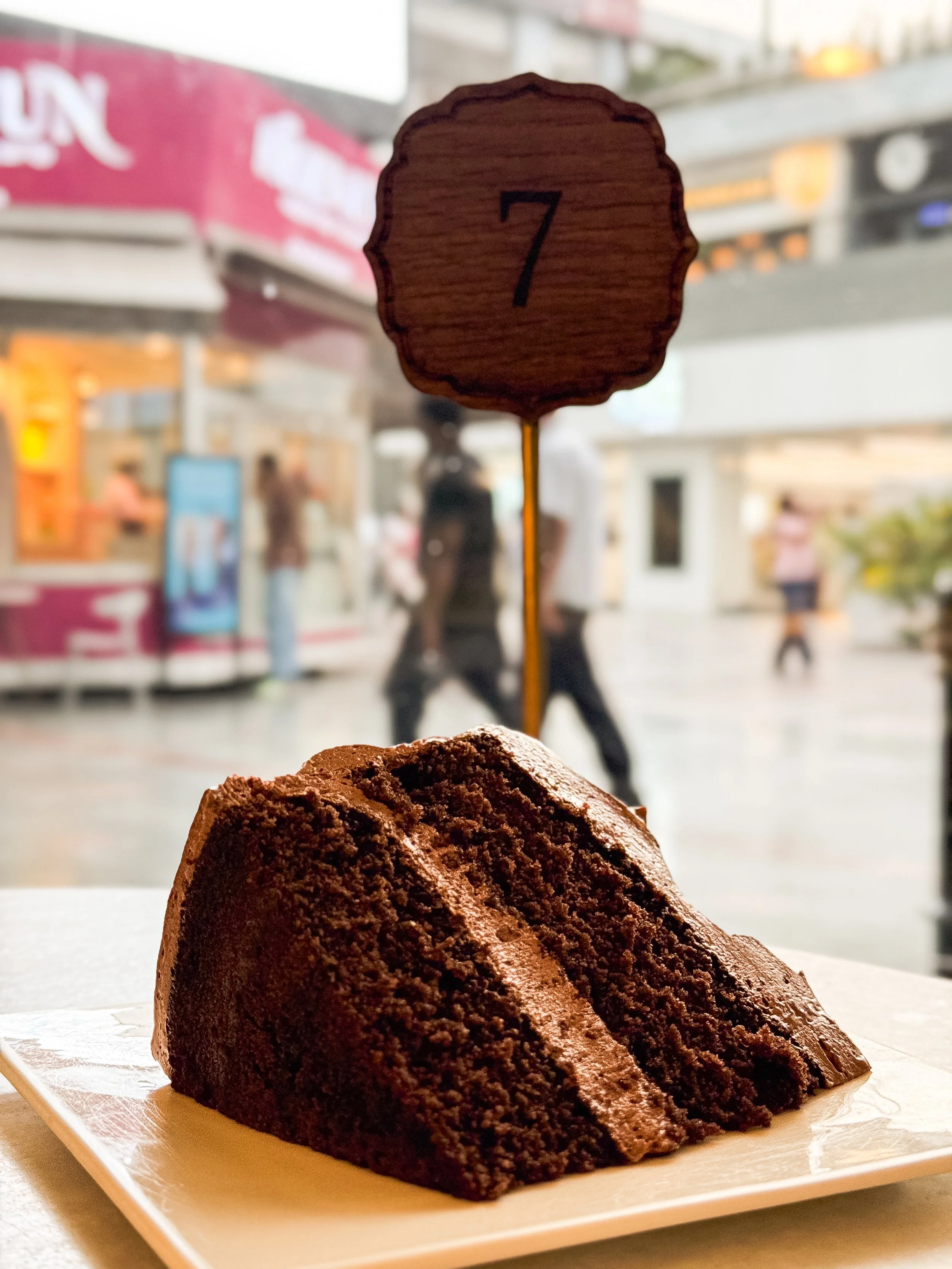 A slice of chocolate cake on a white plate with a sign displaying the number 7 in the background, inside a cafe or bakery, with blurred people and storefronts outside.