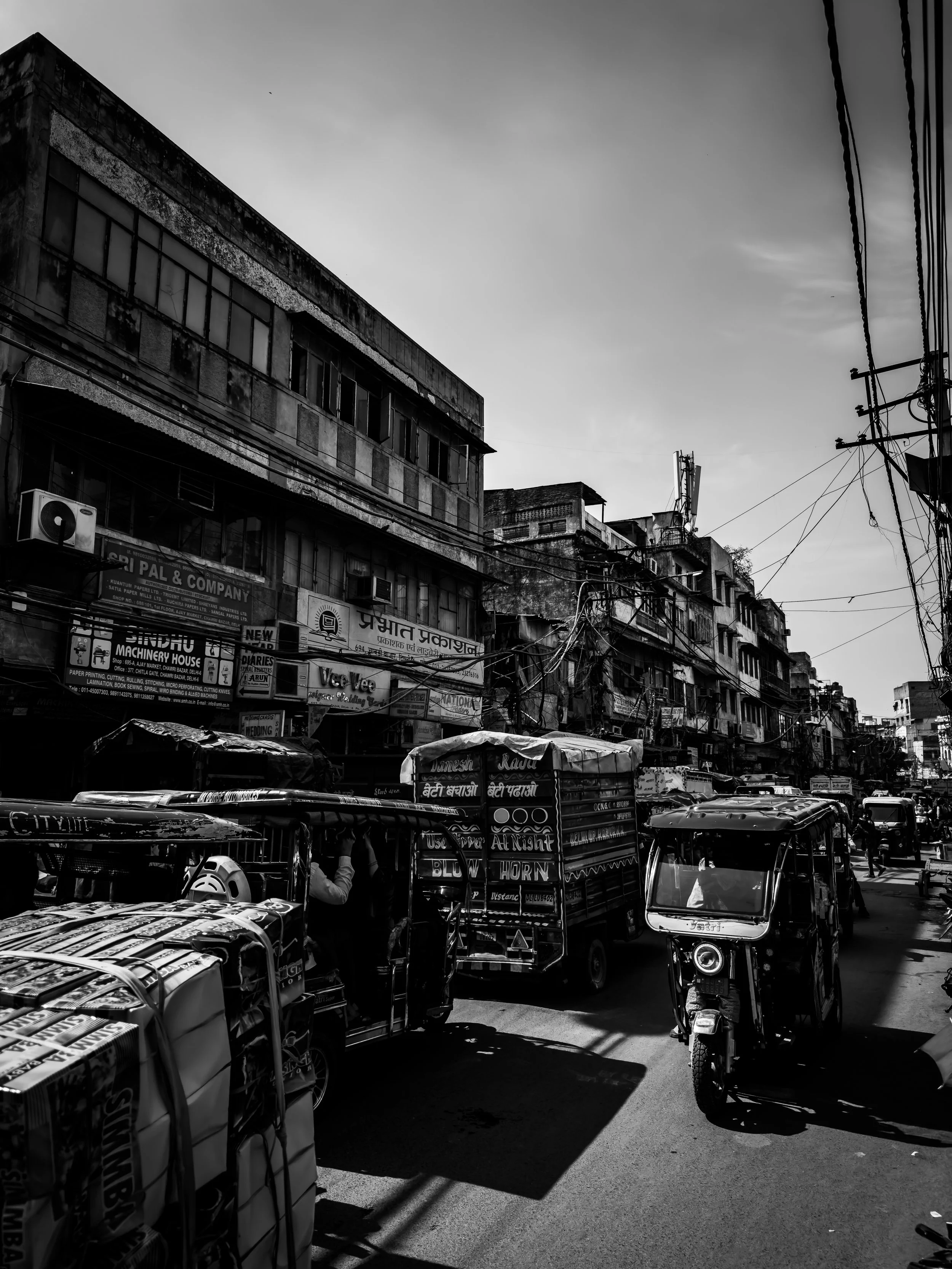 Black and white street scene in an urban area with old buildings, shops, power lines, autorickshaws, and people on the road.
