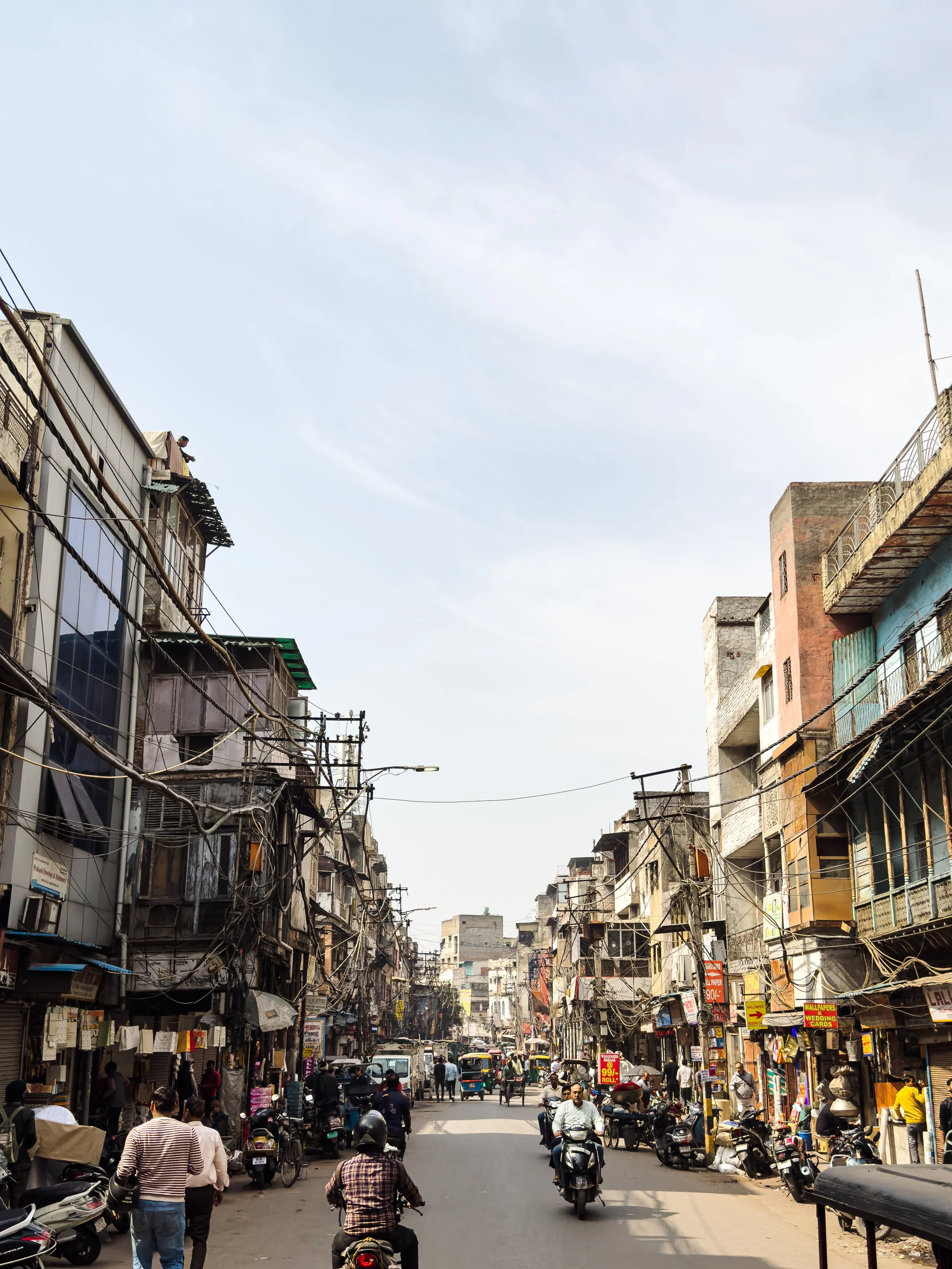 Busy urban street with motorcycles, pedestrians, and small shops on both sides, under a clear blue sky.
