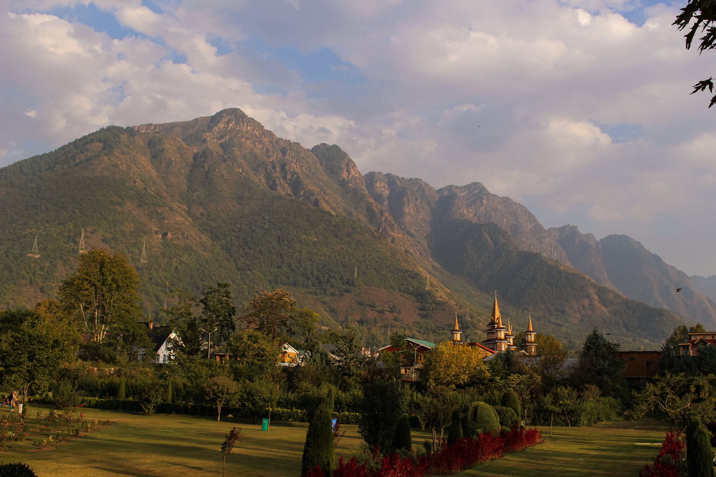 View of a mountainous landscape with a small village and a park in the foreground, featuring trees and lawn, under a partly cloudy sky.