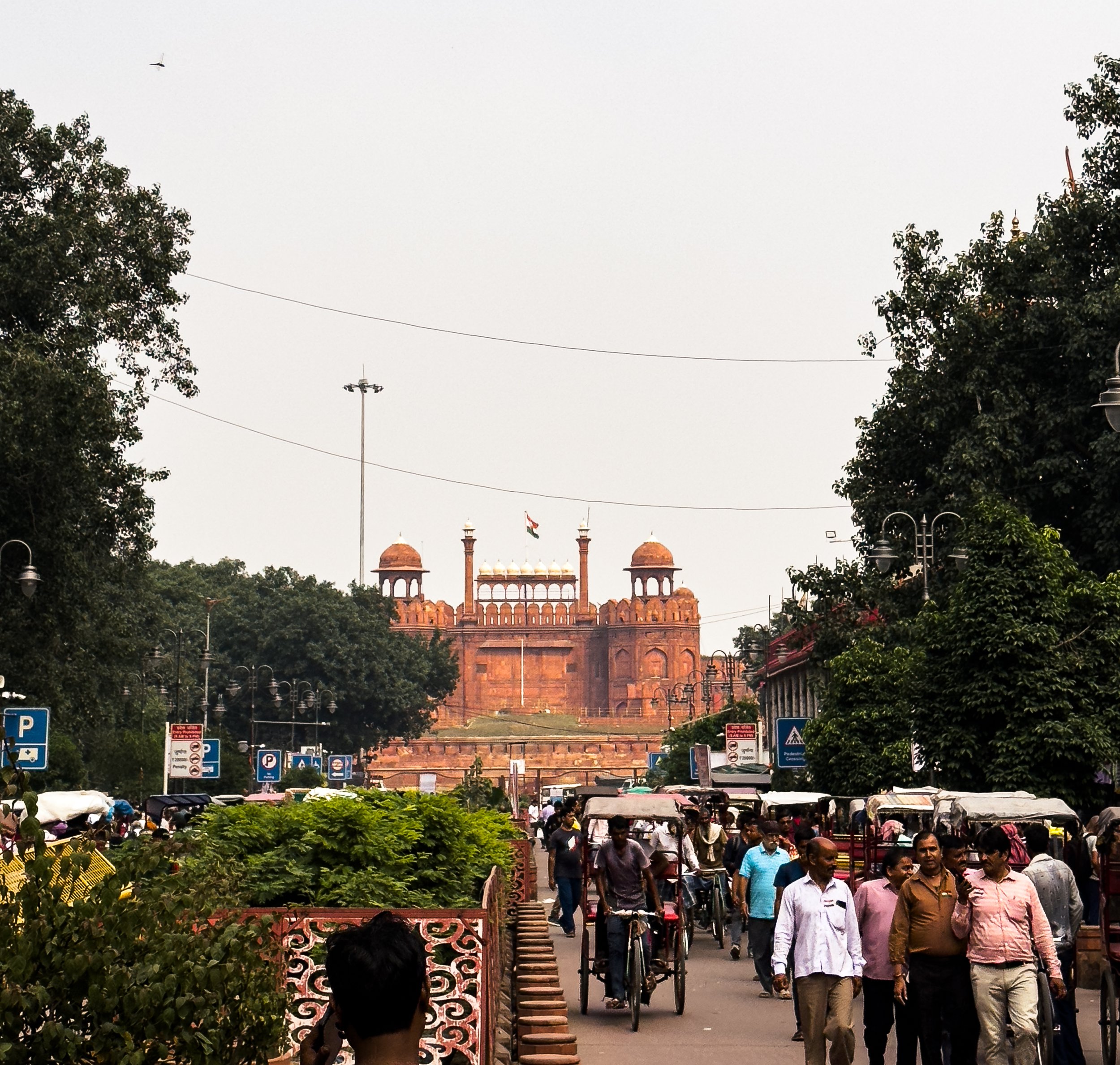 Street scene with people walking and cycling towards Red Fort in Delhi, India, with trees and street lamps on either side.
