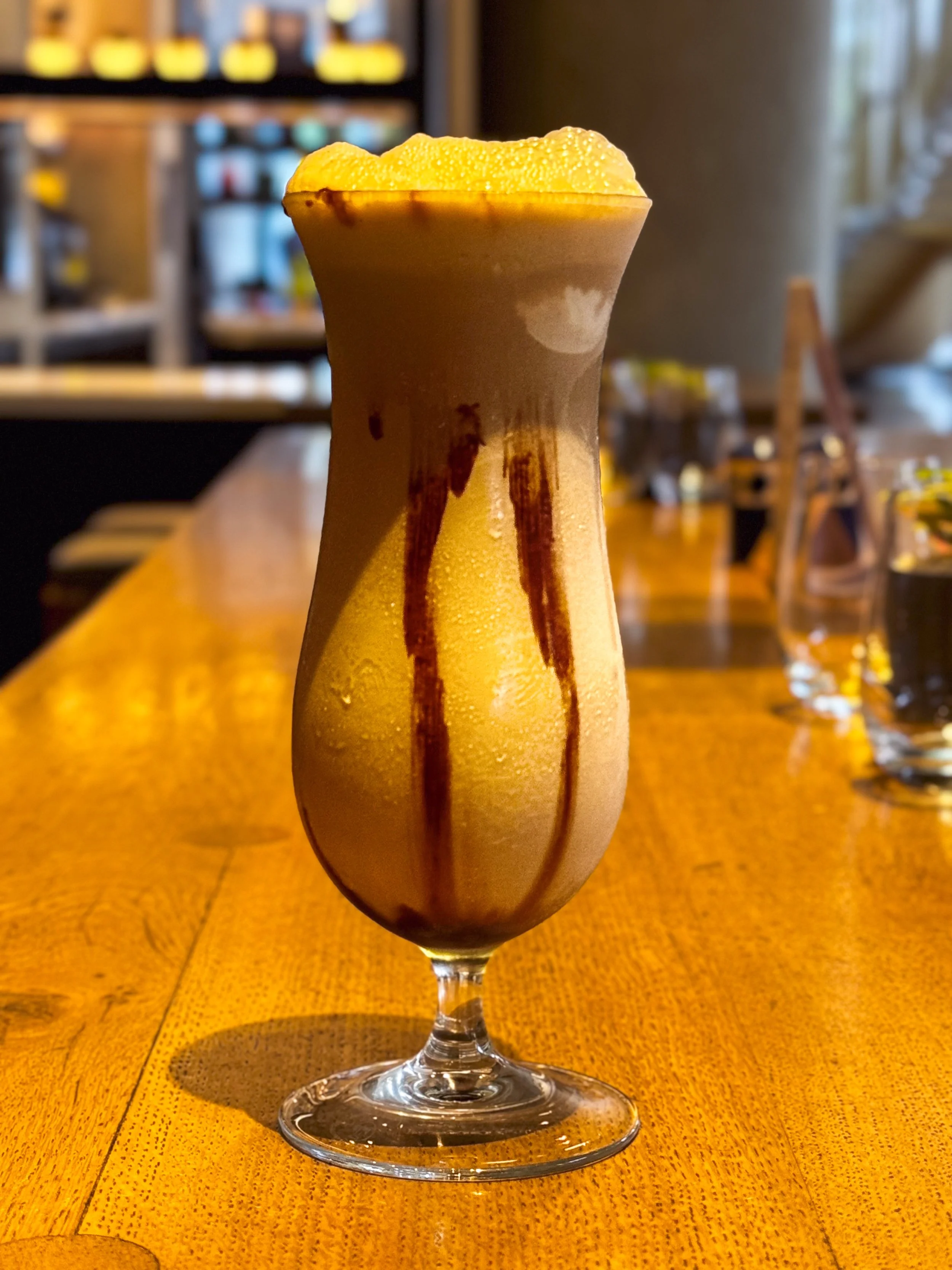 A tall glass filled with a layered iced coffee drink, topped with foam and drizzled with chocolate syrup, on a wooden table in a café.