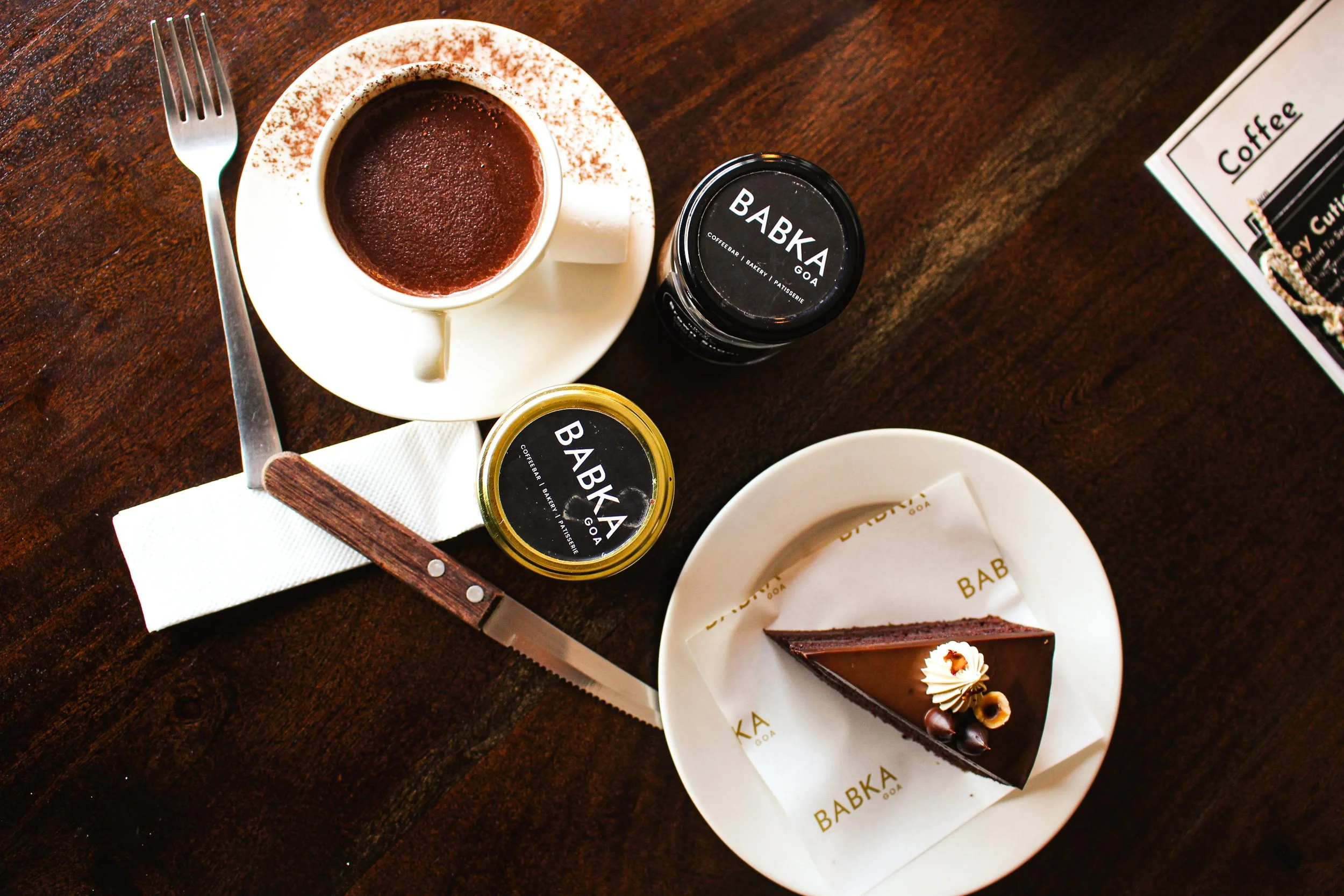 A cup of coffee on a saucer, two jars of Bebka spread, a slice of chocolate cake with decorations on a plate, a fork, a bread knife, a napkin, and part of a menu on a dark wooden table.