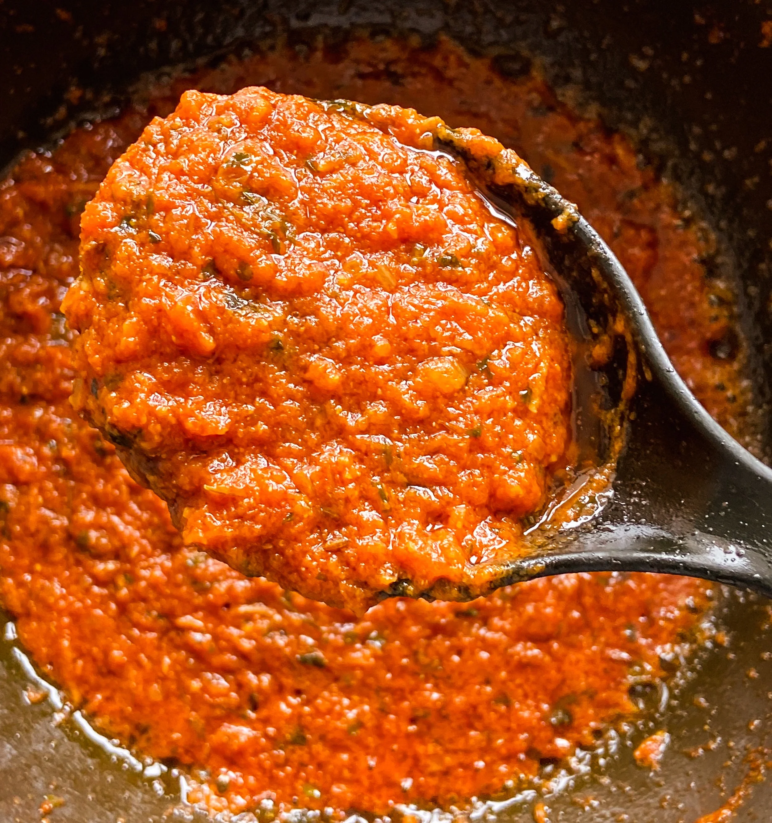 A close-up of a spoonful of thick, red tomato sauce being lifted from a pot, with the sauce's textured, chunky appearance visible.