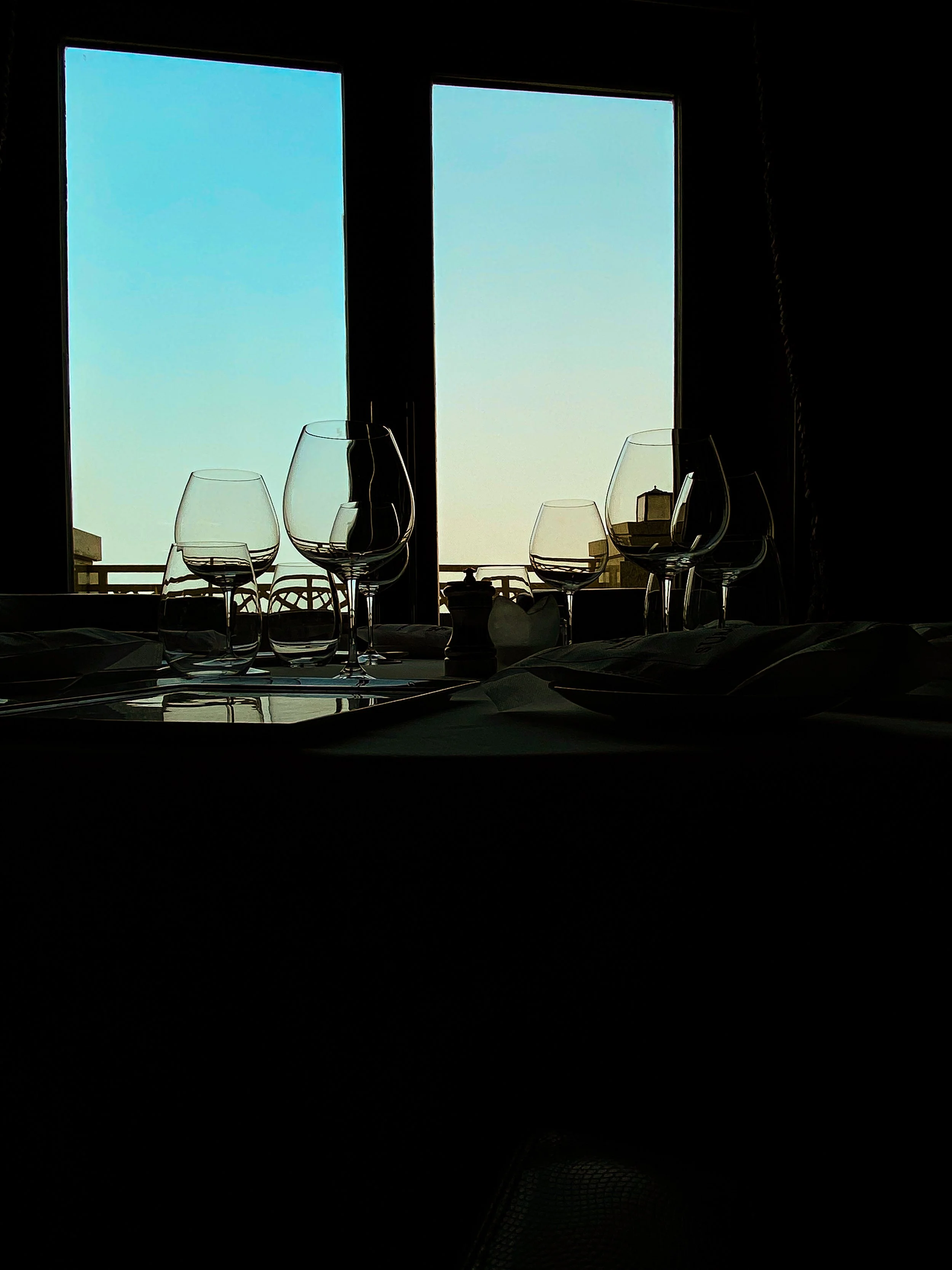 Silhouetted table with empty wine glasses and a pepper shaker in front of a window showing a clear sky.
