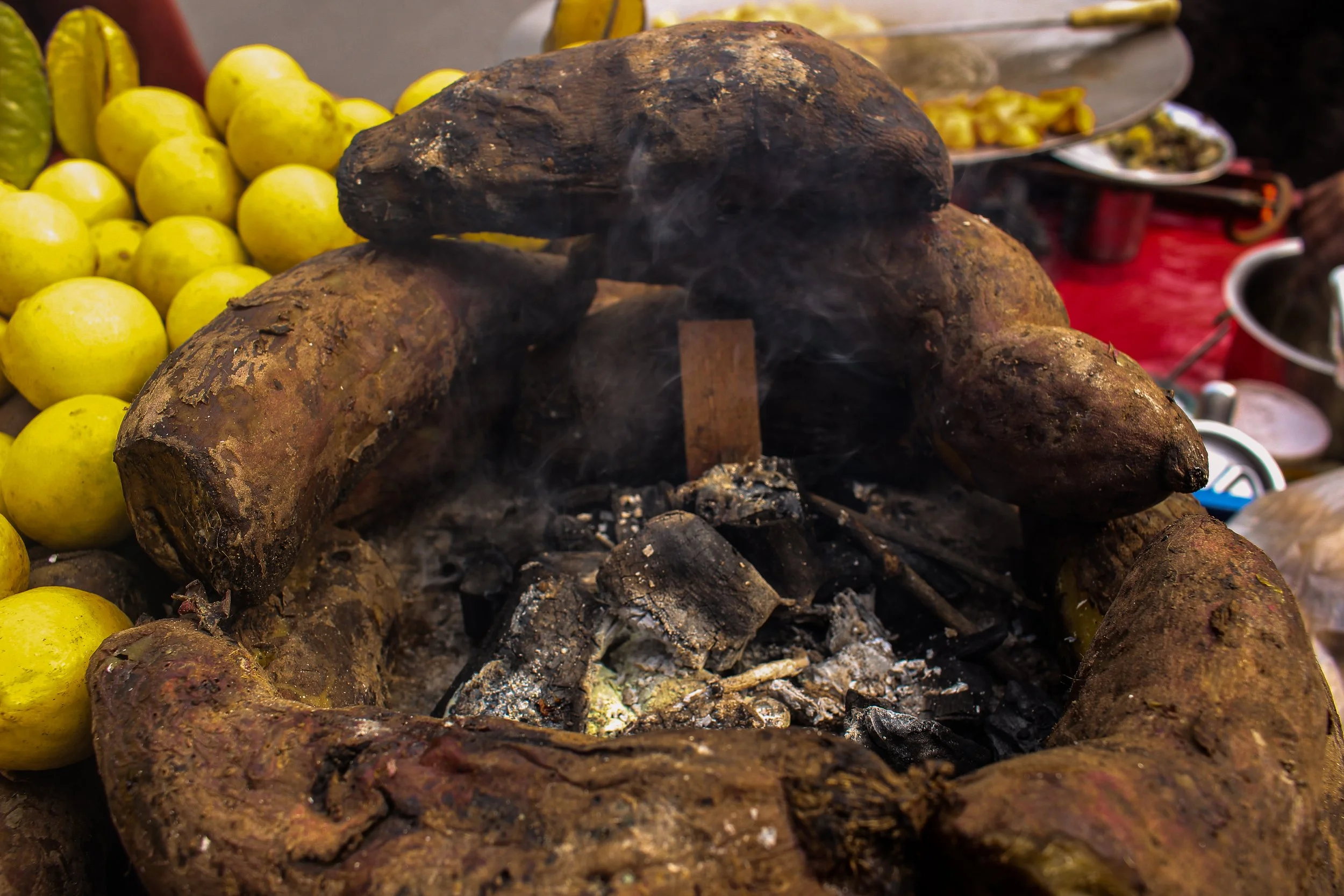 A traditional clay stove with burning charcoal, surrounded by roasted sweet potatoes, with yellow fruits and some bowls in the background.