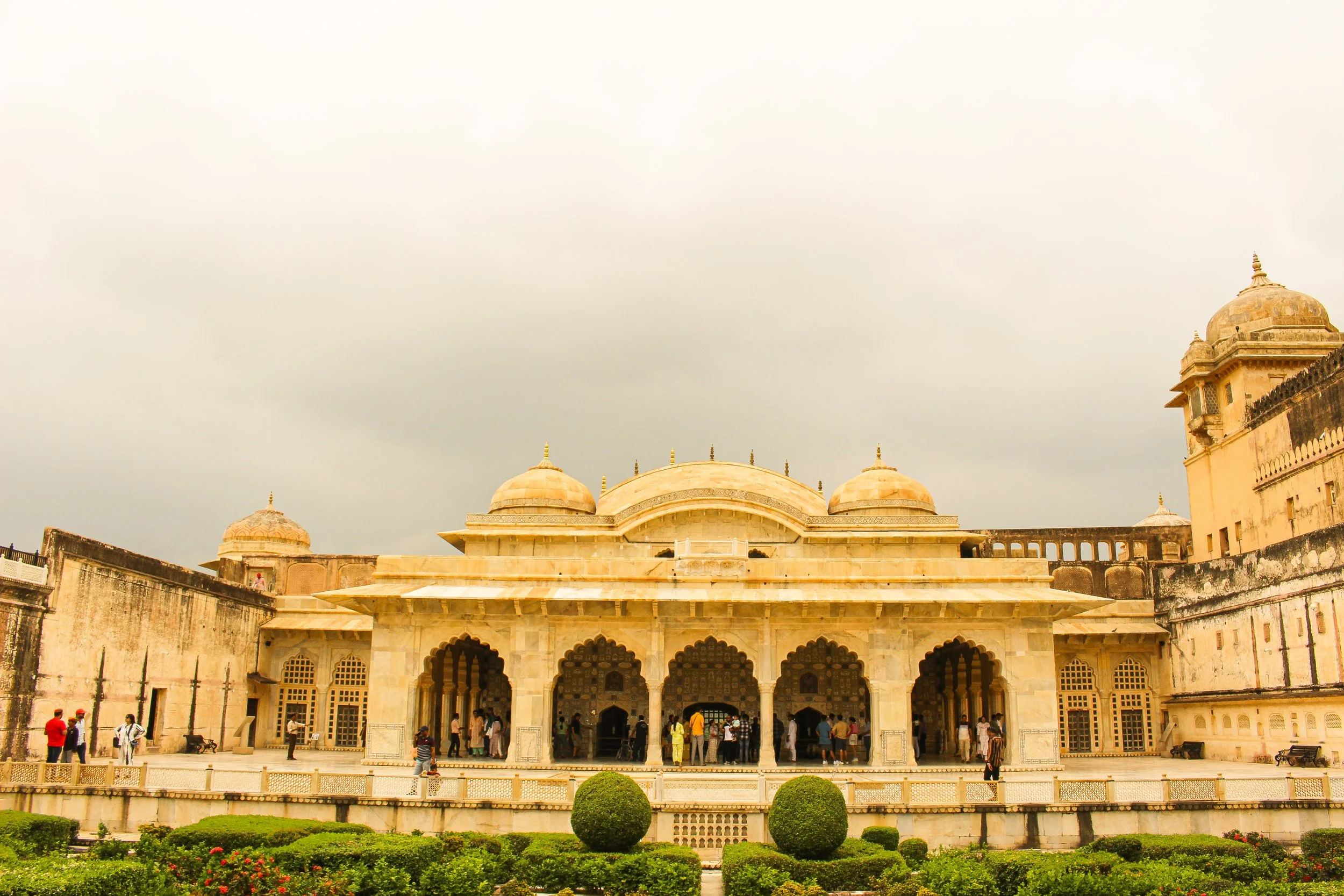 Historical building with domes and arched entrances, surrounded by a garden with manicured bushes and people walking around, under an overcast sky.