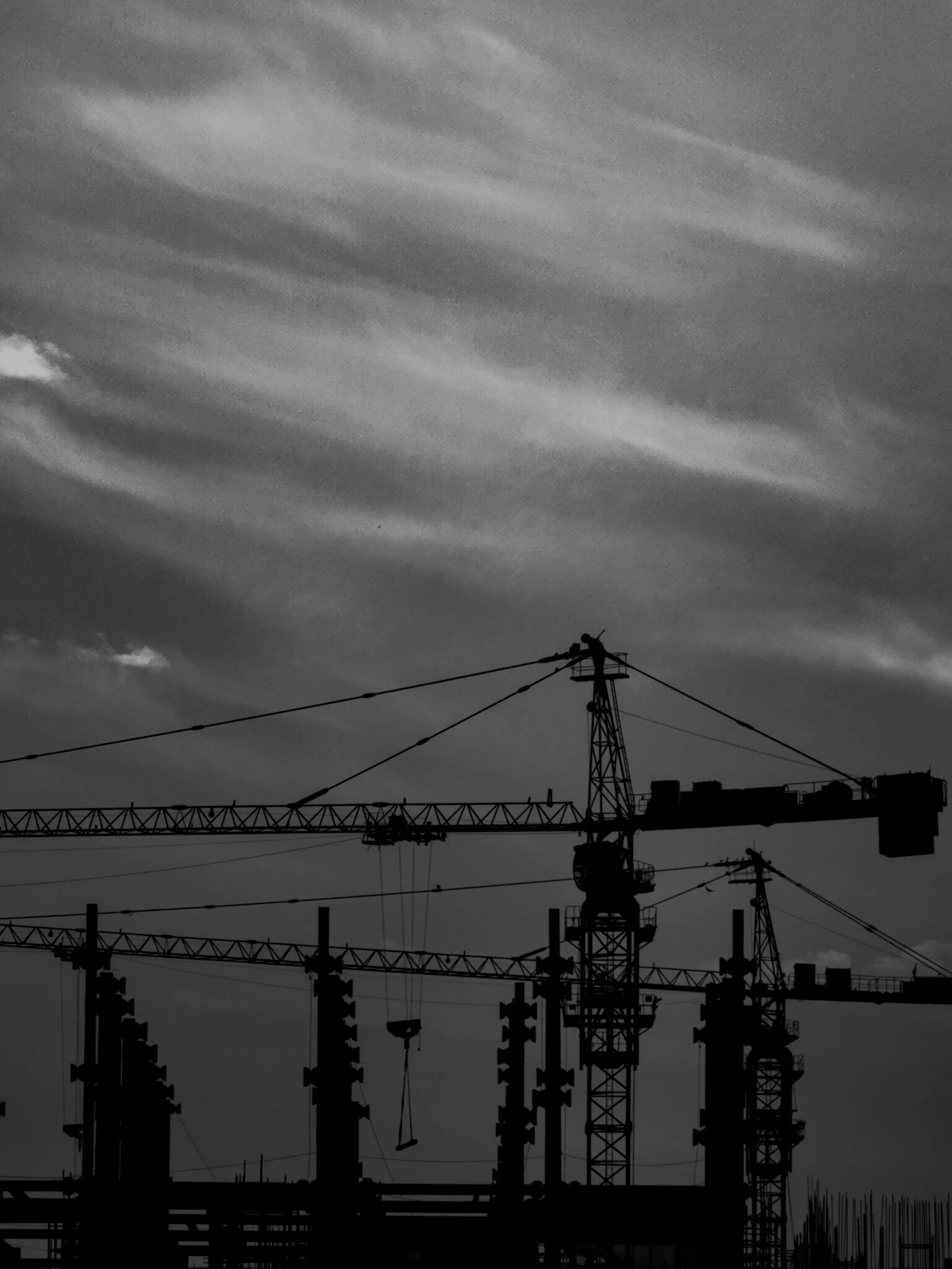 Silhouettes of construction cranes and building structures against a cloudy sky at dusk or dawn.