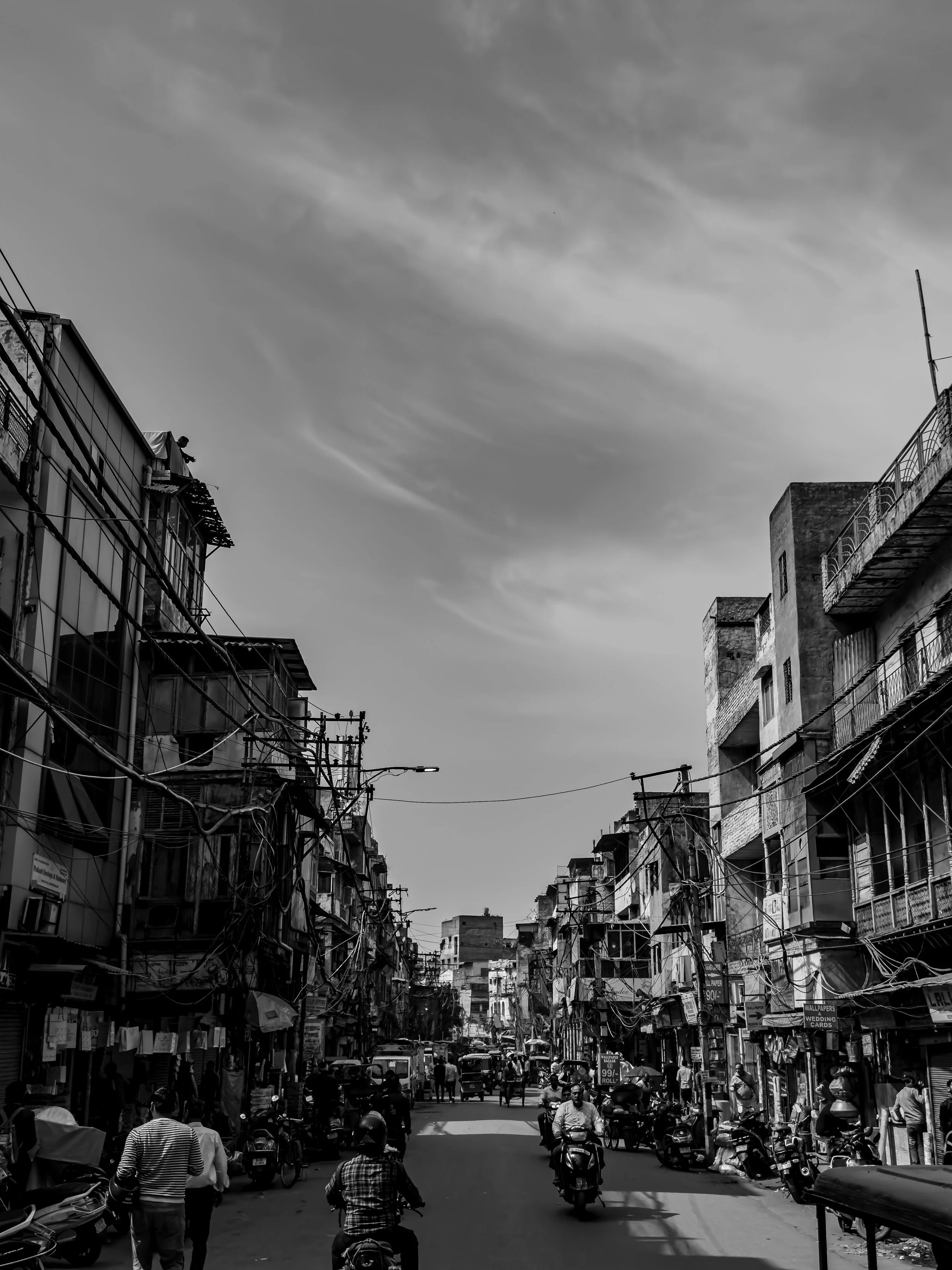 A busy urban street scene with buildings on both sides, numerous electrical wires overhead, and people walking or riding motorbikes on the street.