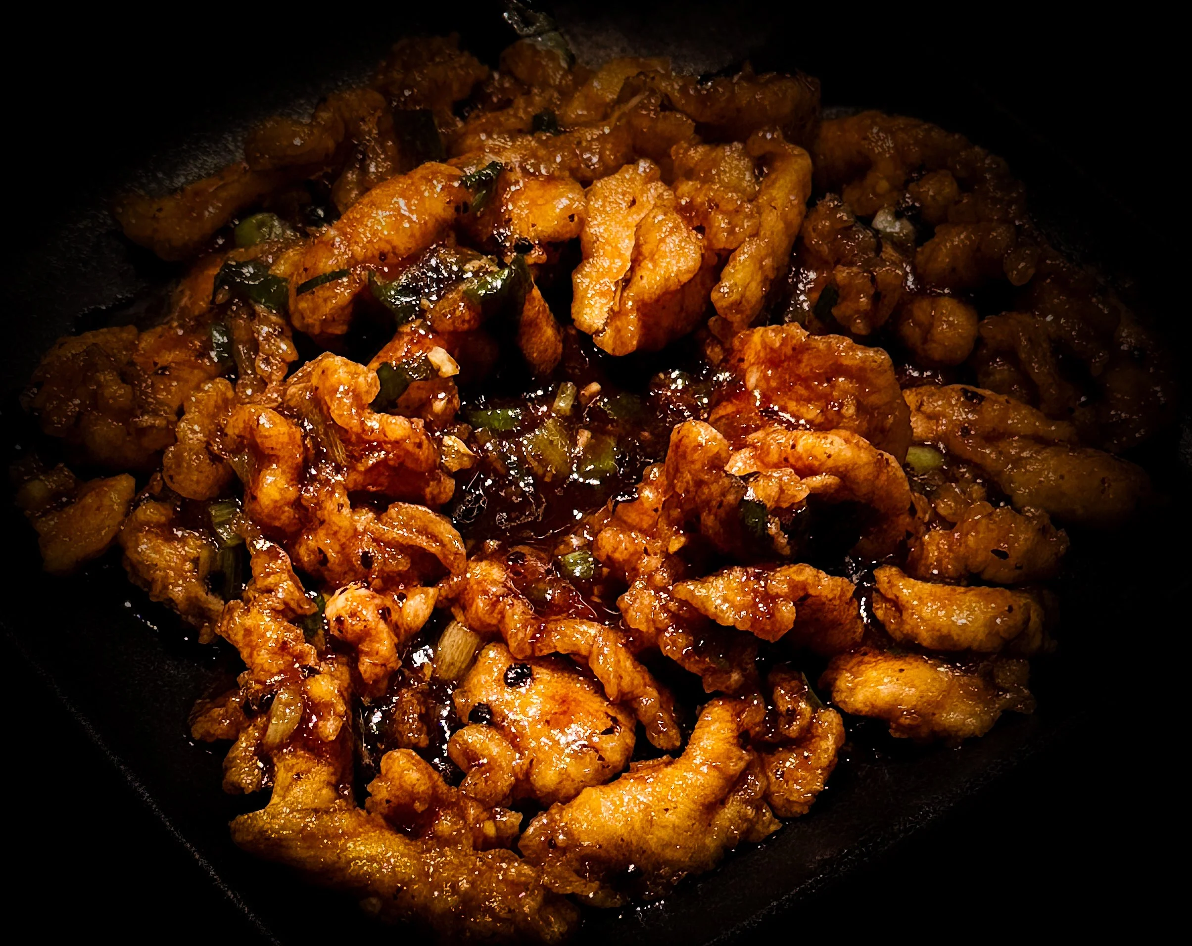 Close-up of stir-fried chicken with sauce and green scallions in a black skillet.