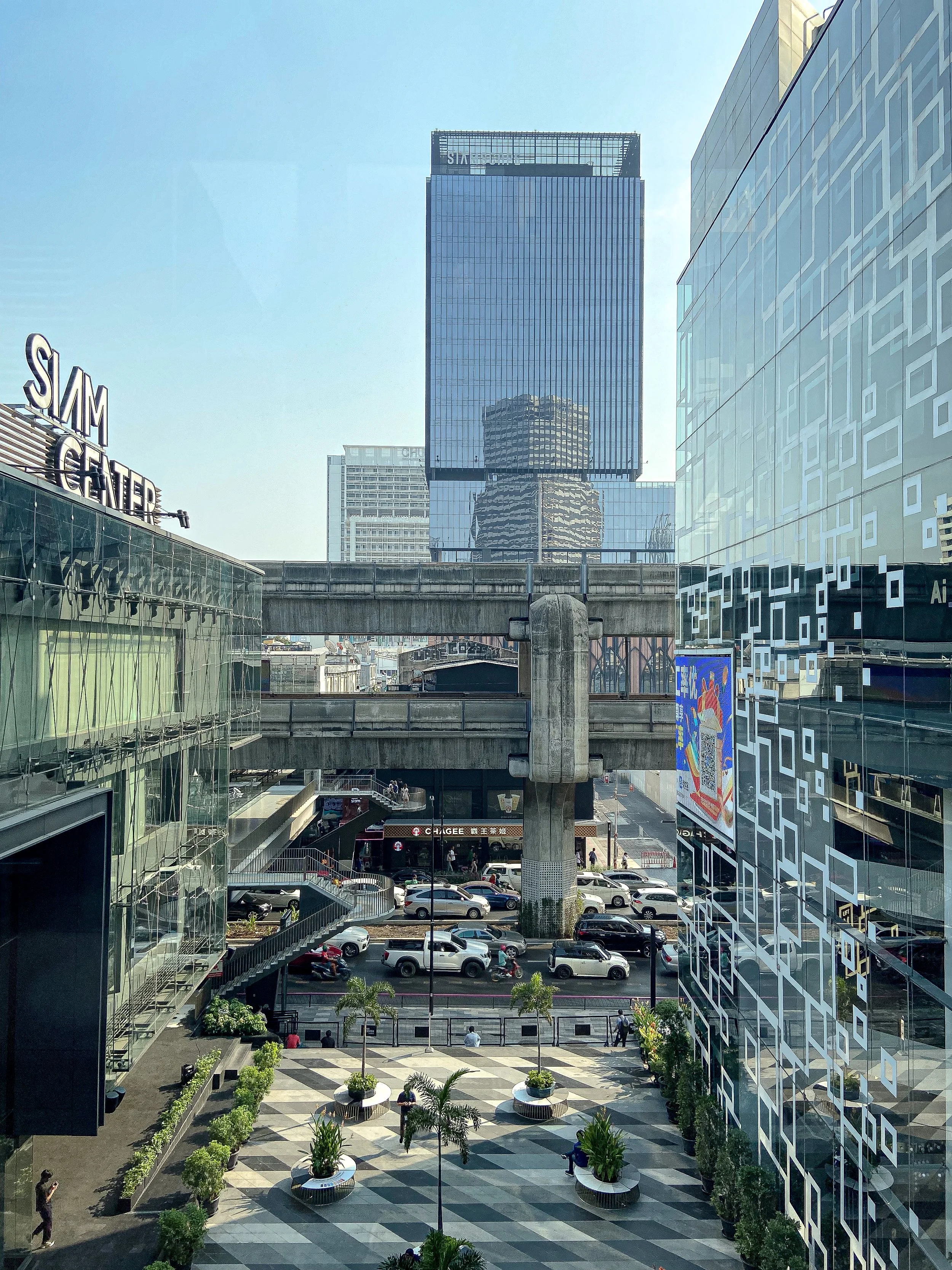 Cityscape view showing modern high-rise buildings, a pedestrian plaza with potted plants, and a busy street with cars and people.