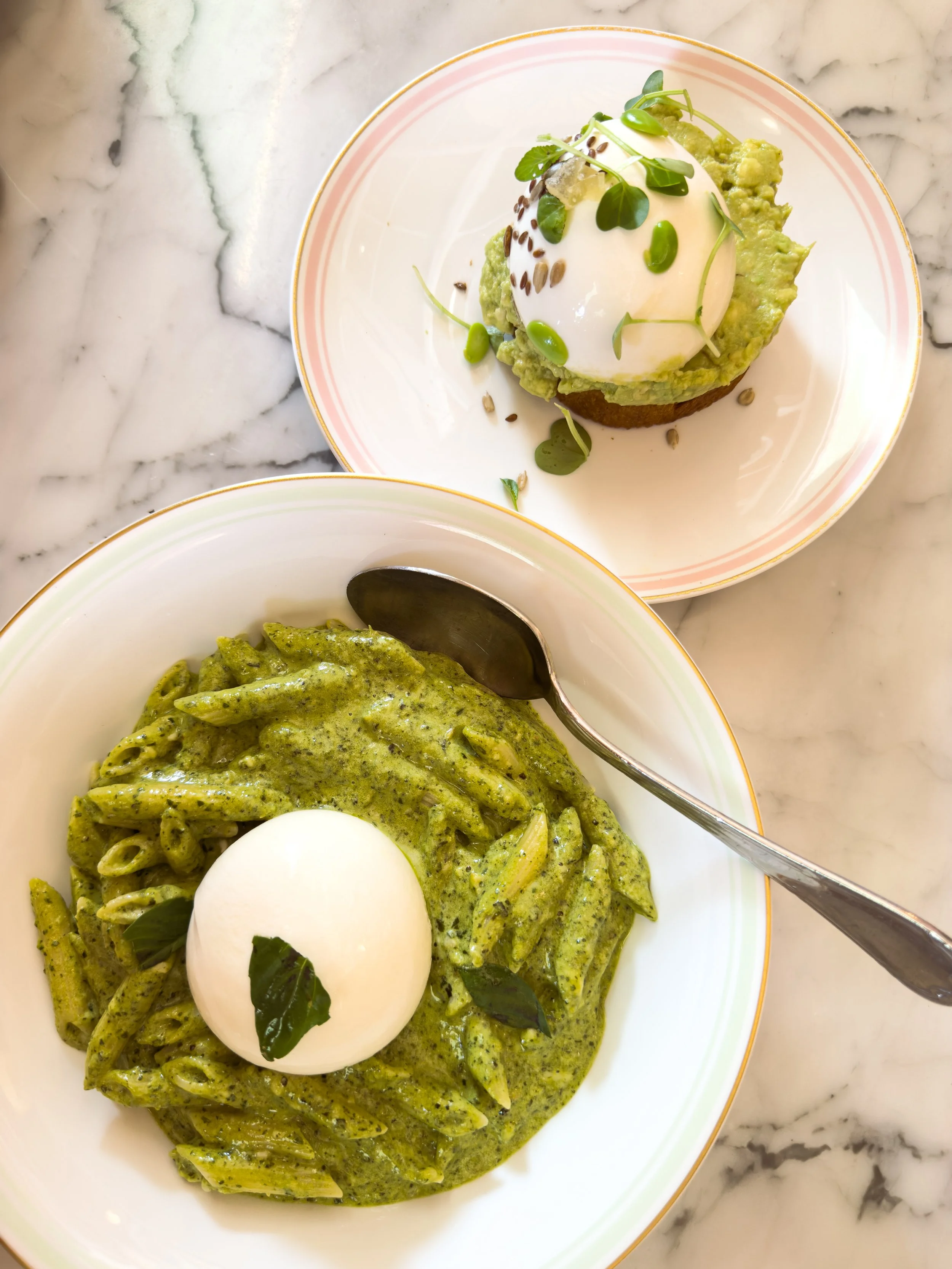 A bowl of green pesto pasta topped with a poached egg and fresh herbs and a plate with avocado toast topped with a poached egg and microgreens, all served on a marble table.