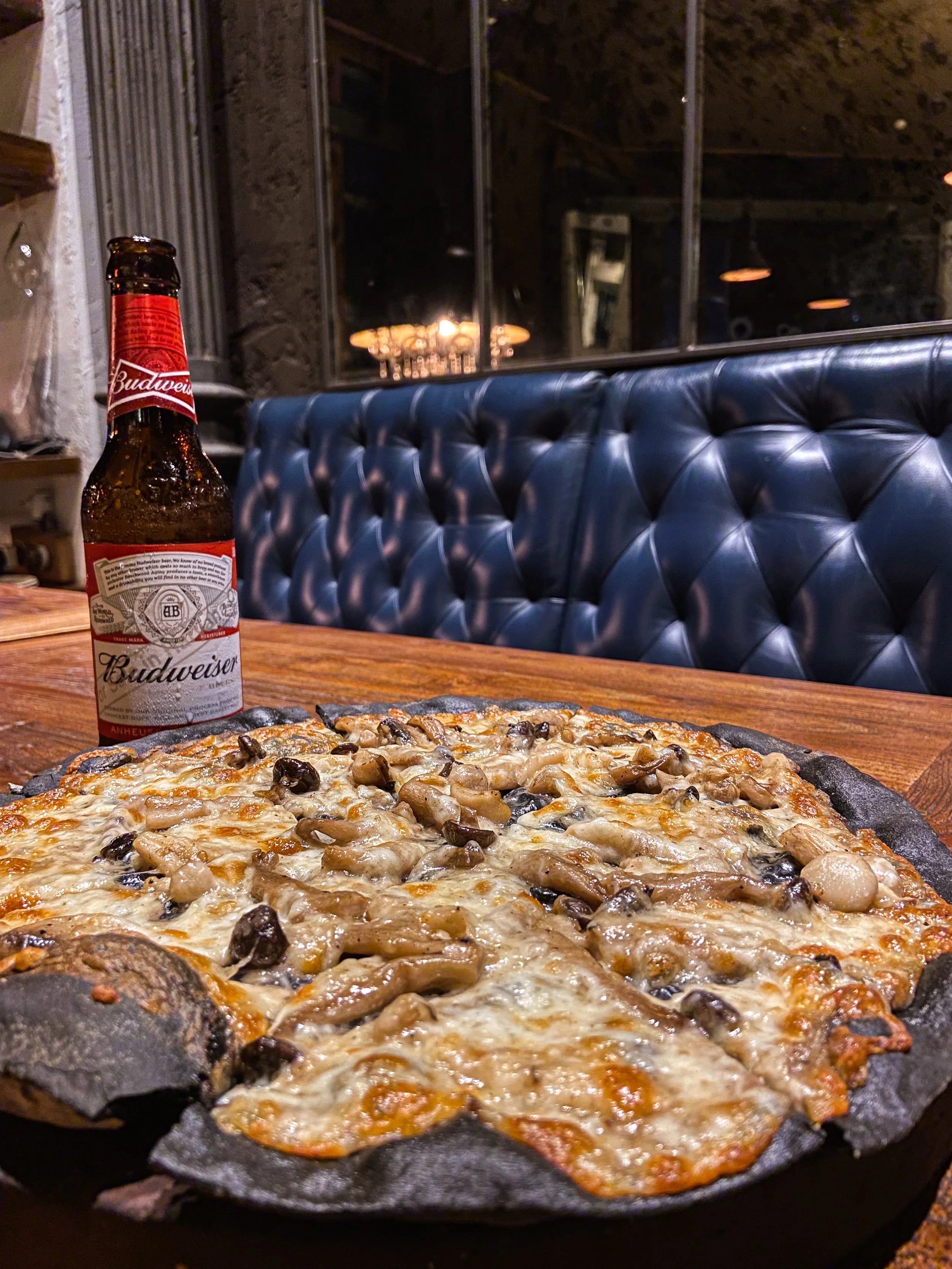 A pizza with mushrooms and cheese on a table with a bottle of Budweiser beer in a restaurant setting.