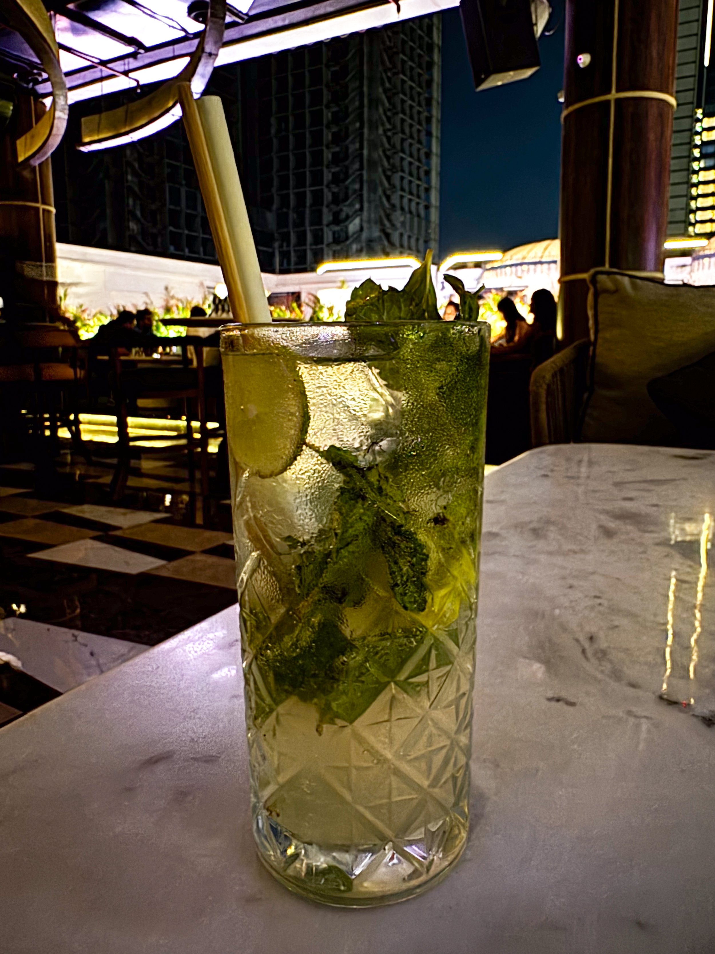 A tall glass of mojito with mint leaves, lime slices, ice cubes, and a straw on a marble table in a restaurant or bar with a dark interior and some people in the background.