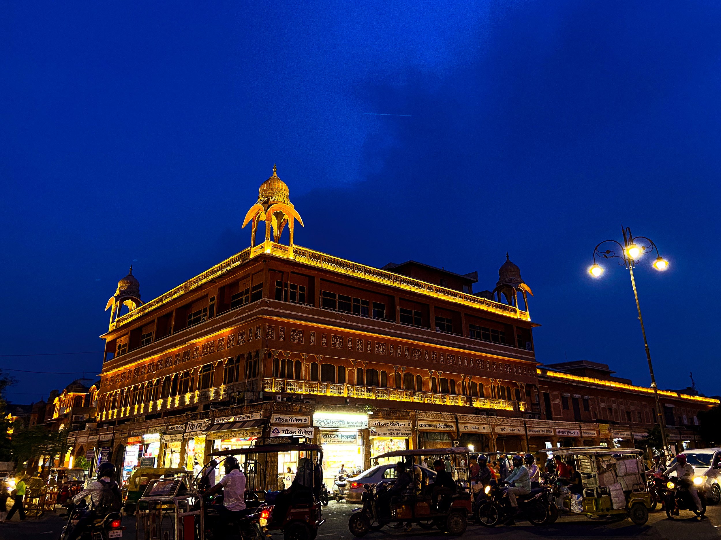A brightly lit multi-story building at night with traditional architecture, multiple shops on the ground floor, and a busy street with motorcycles and cars in front.