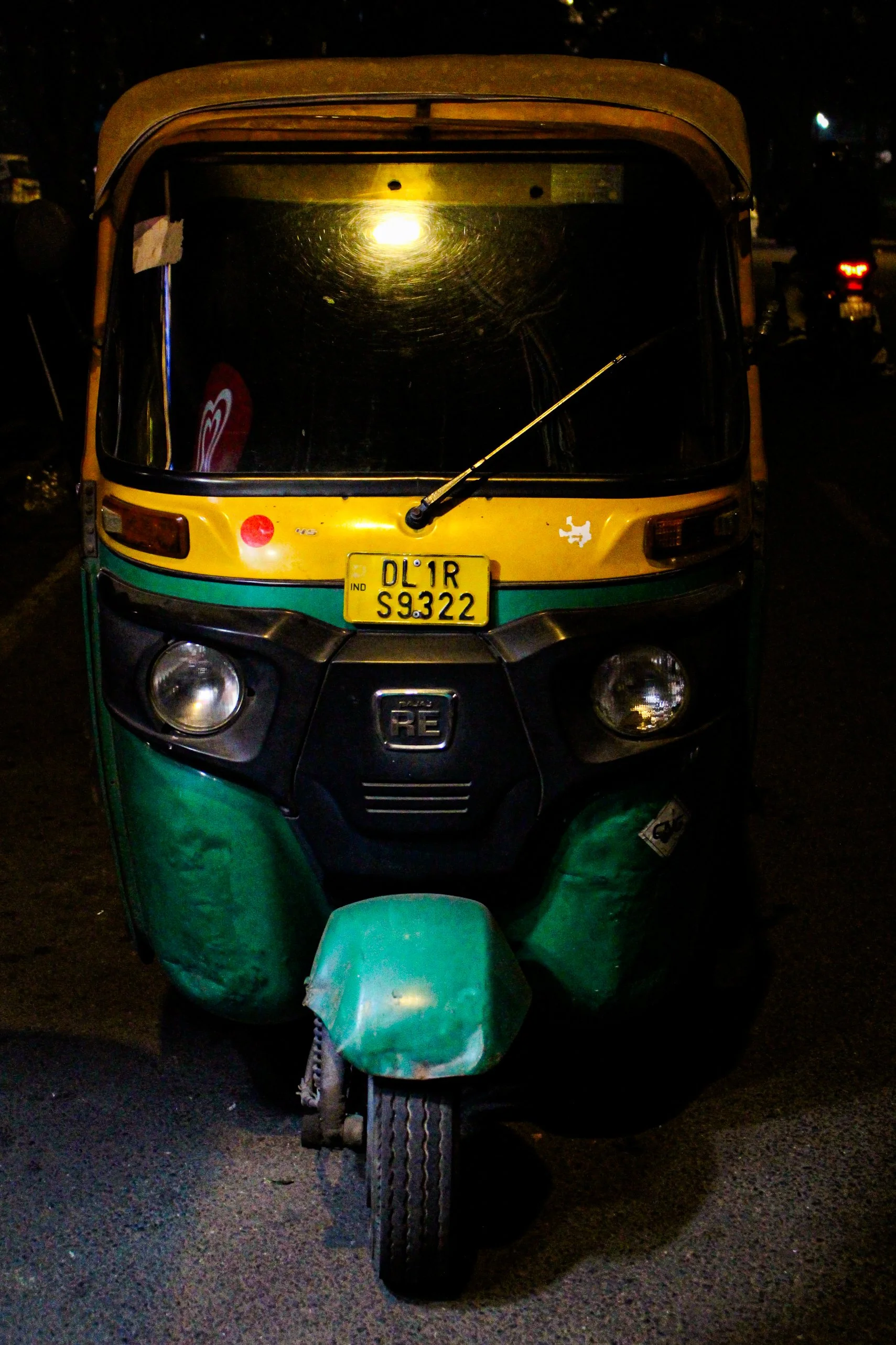 Front view of a yellow and green auto rickshaw at night, with a yellow license plate reading DL 1R S 9322 and a dark background.
