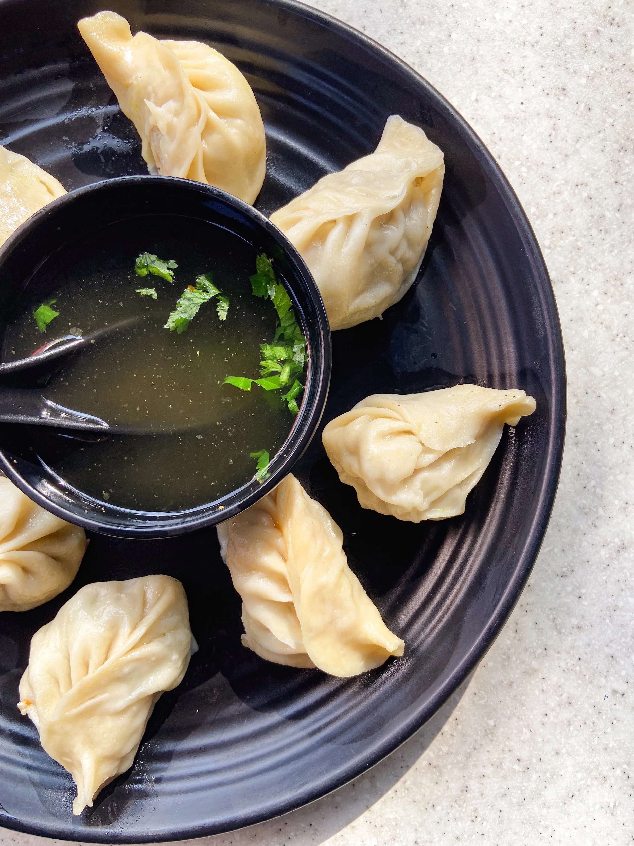 A black plate with six steamed dumplings and a small black bowl of dipping sauce garnished with cilantro, on a light-colored countertop.