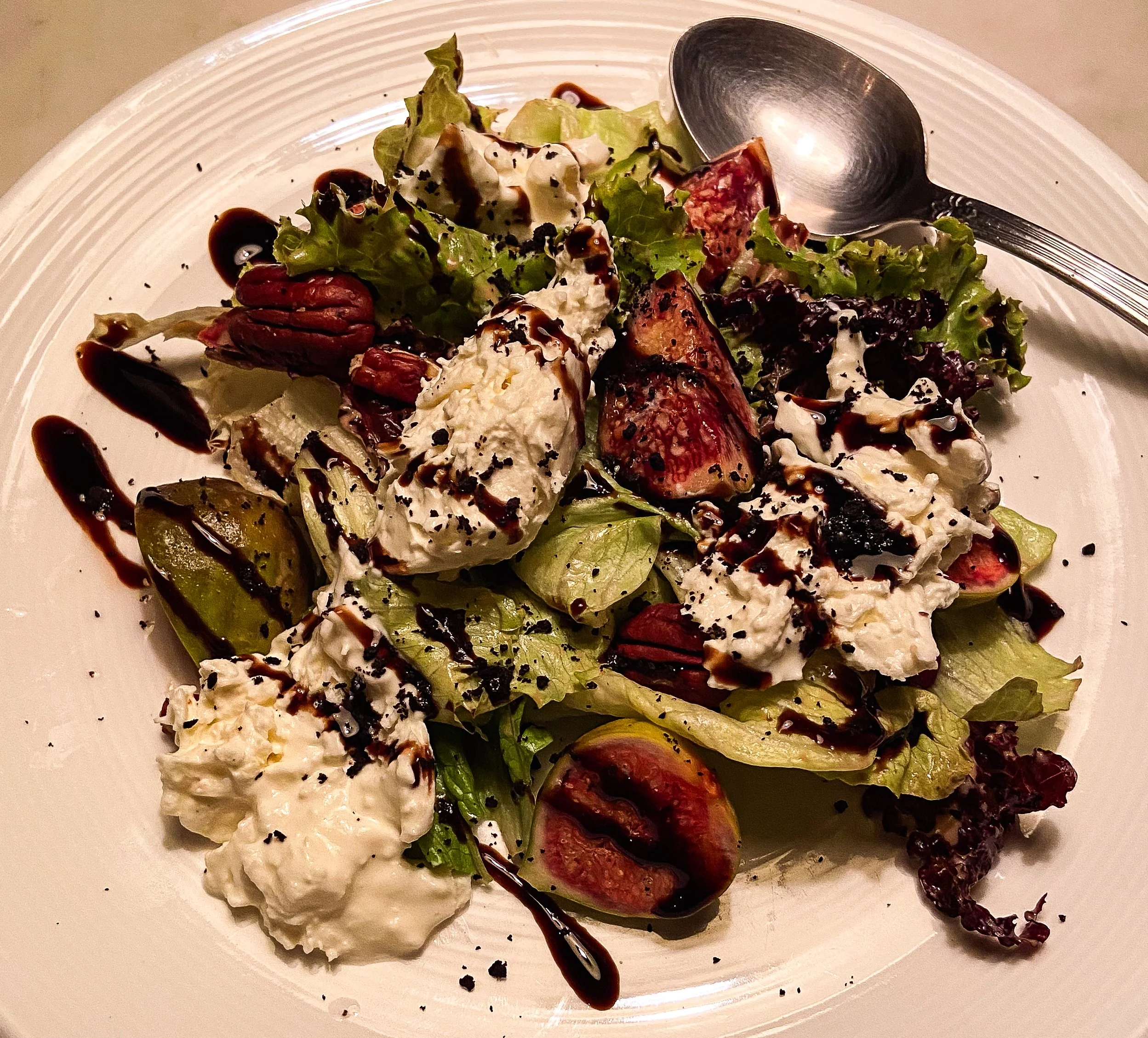 A salad with lettuce, strawberries, pecans, and cheese, drizzled with balsamic glaze on a white plate with a silver spoon.