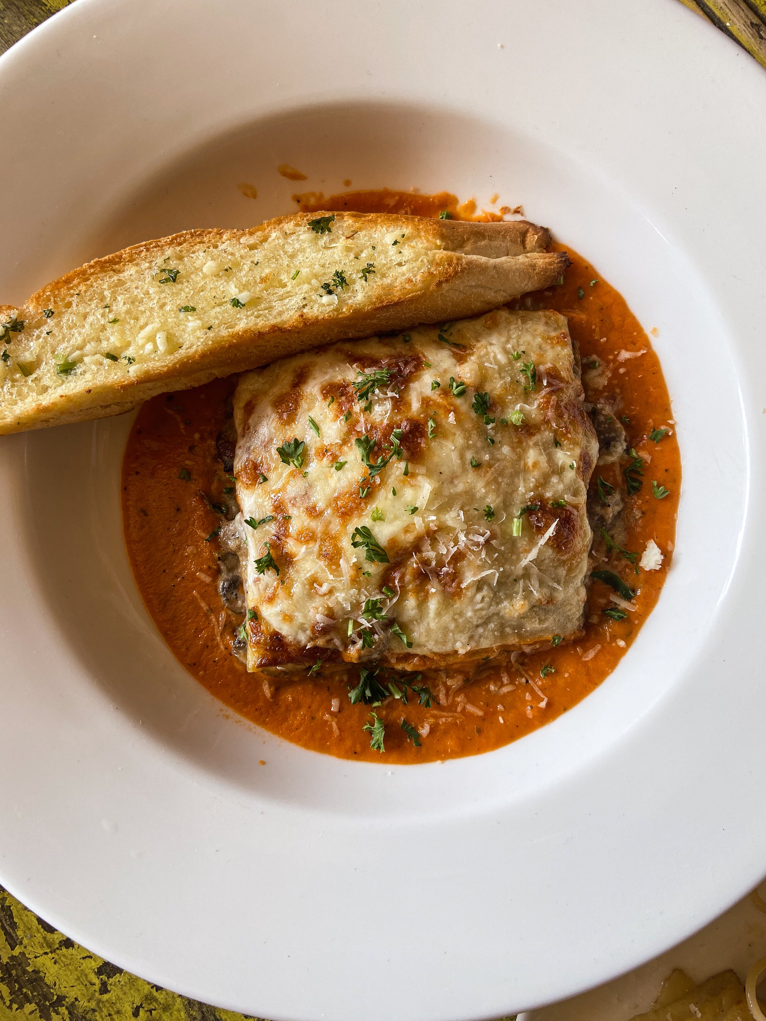 Baked stuffed chicken breast with cheese and herbs, covered in tomato sauce, served with a slice of garlic bread, garnished with chopped parsley on a white plate.