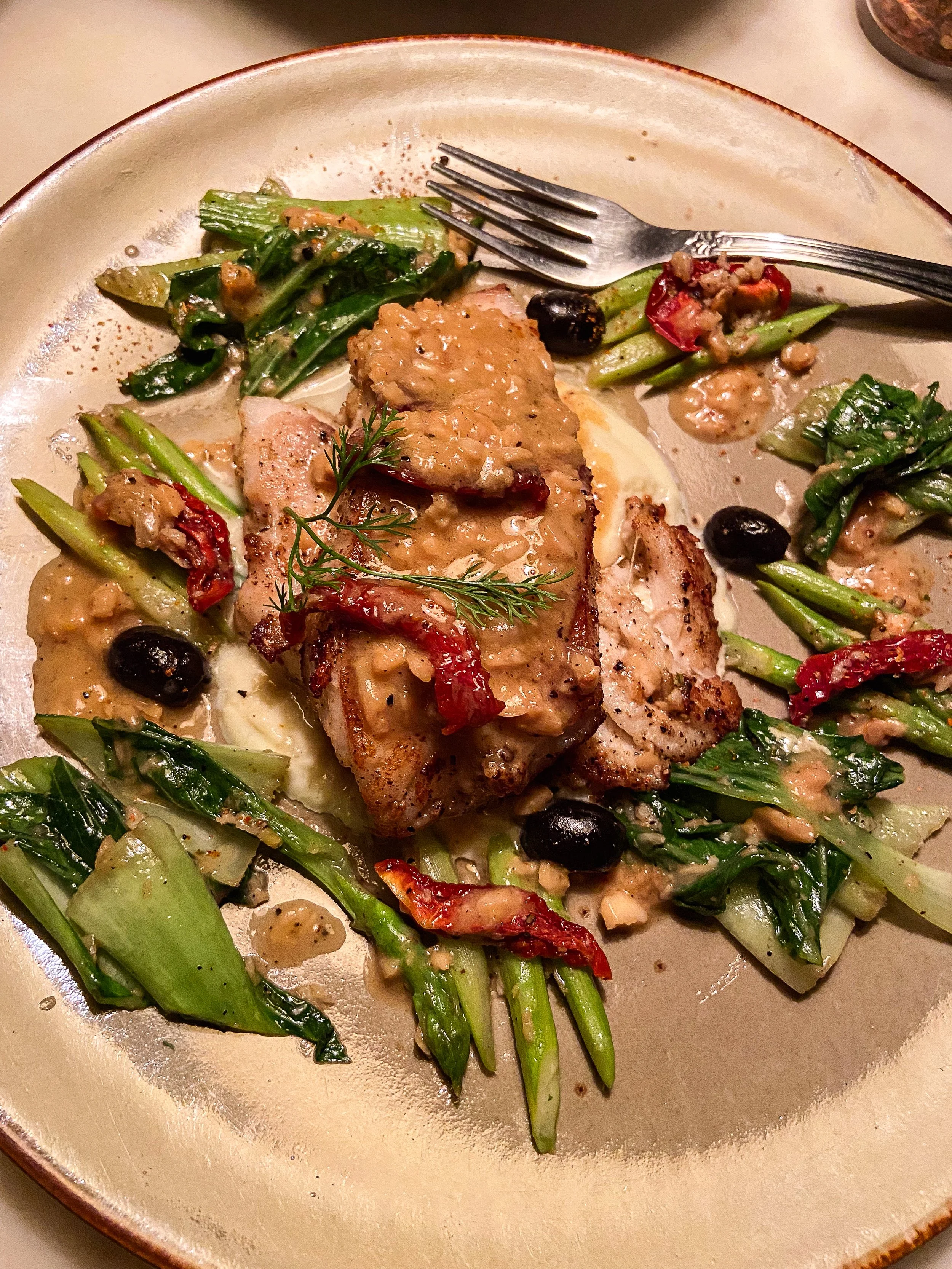 Plate with cooked chicken topped with sauce, surrounded by sautéed greens, red peppers, black olives, and drizzled with sauce.