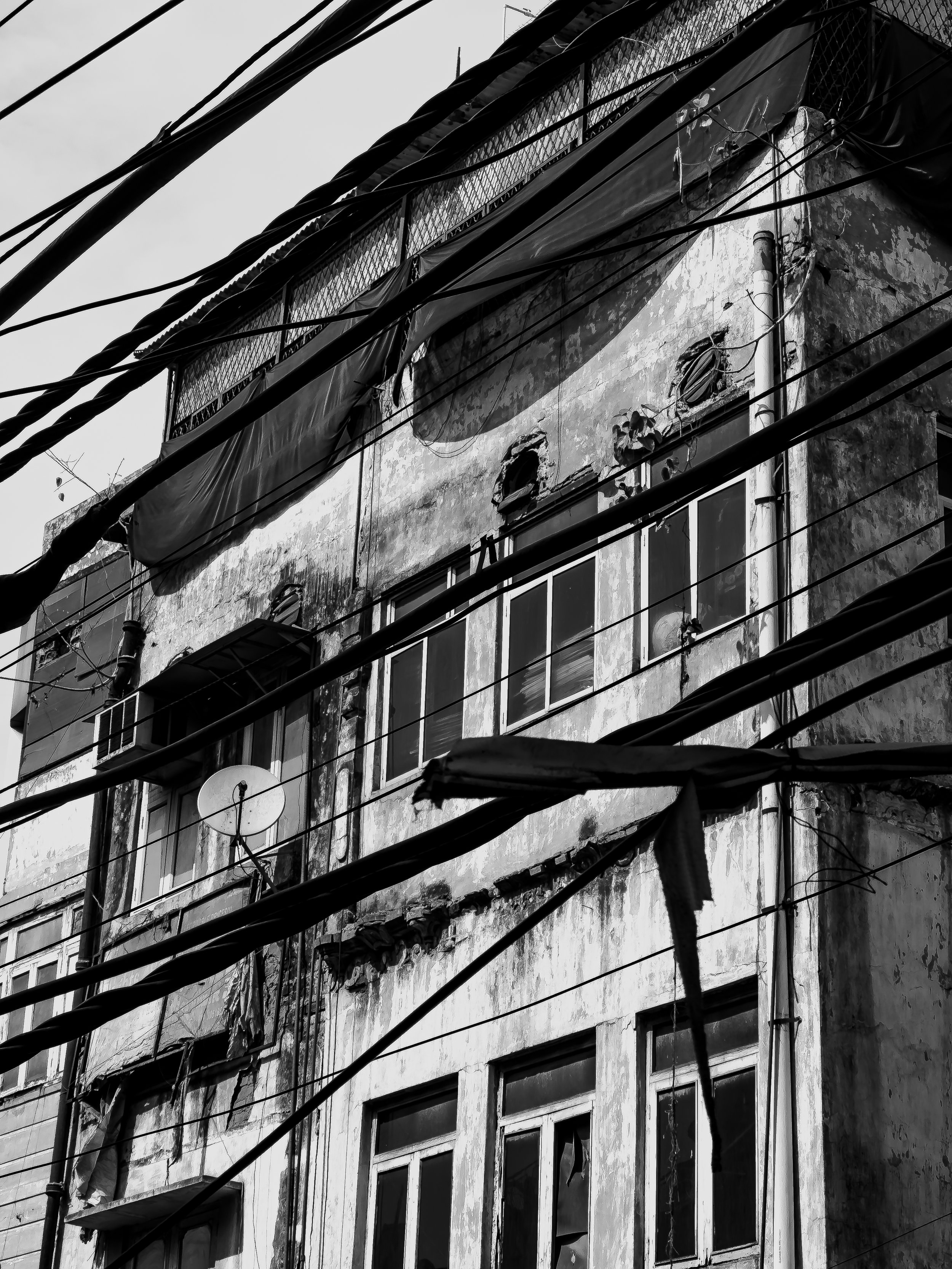 Black and white photo of an old, weathered apartment building with broken windows, dirt, and damaged walls, with tangled electrical wires in the foreground.