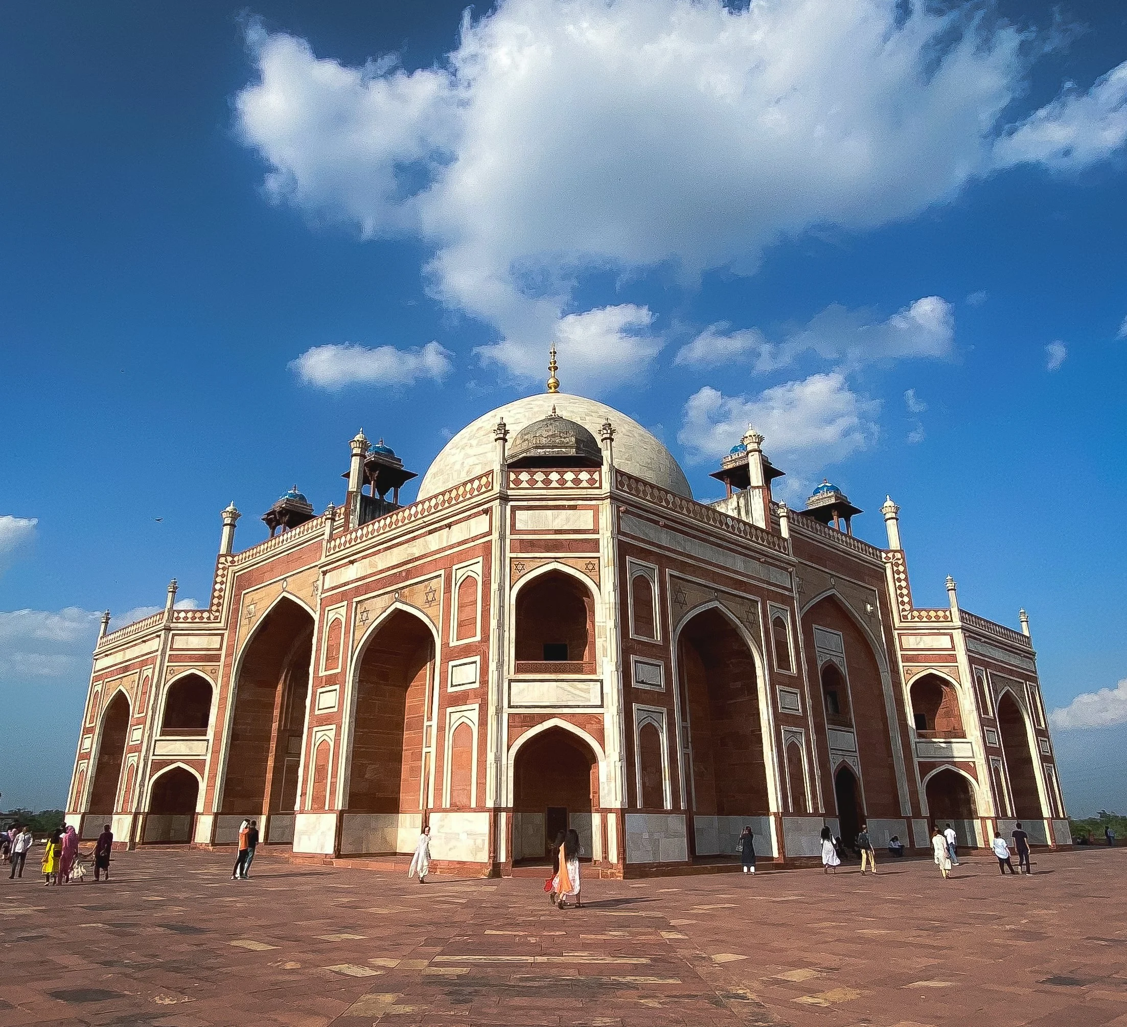 A large red sandstone building with white marble accents and a central white dome, surrounded by smaller domes and arches, under a partly cloudy blue sky, with people walking around its base.