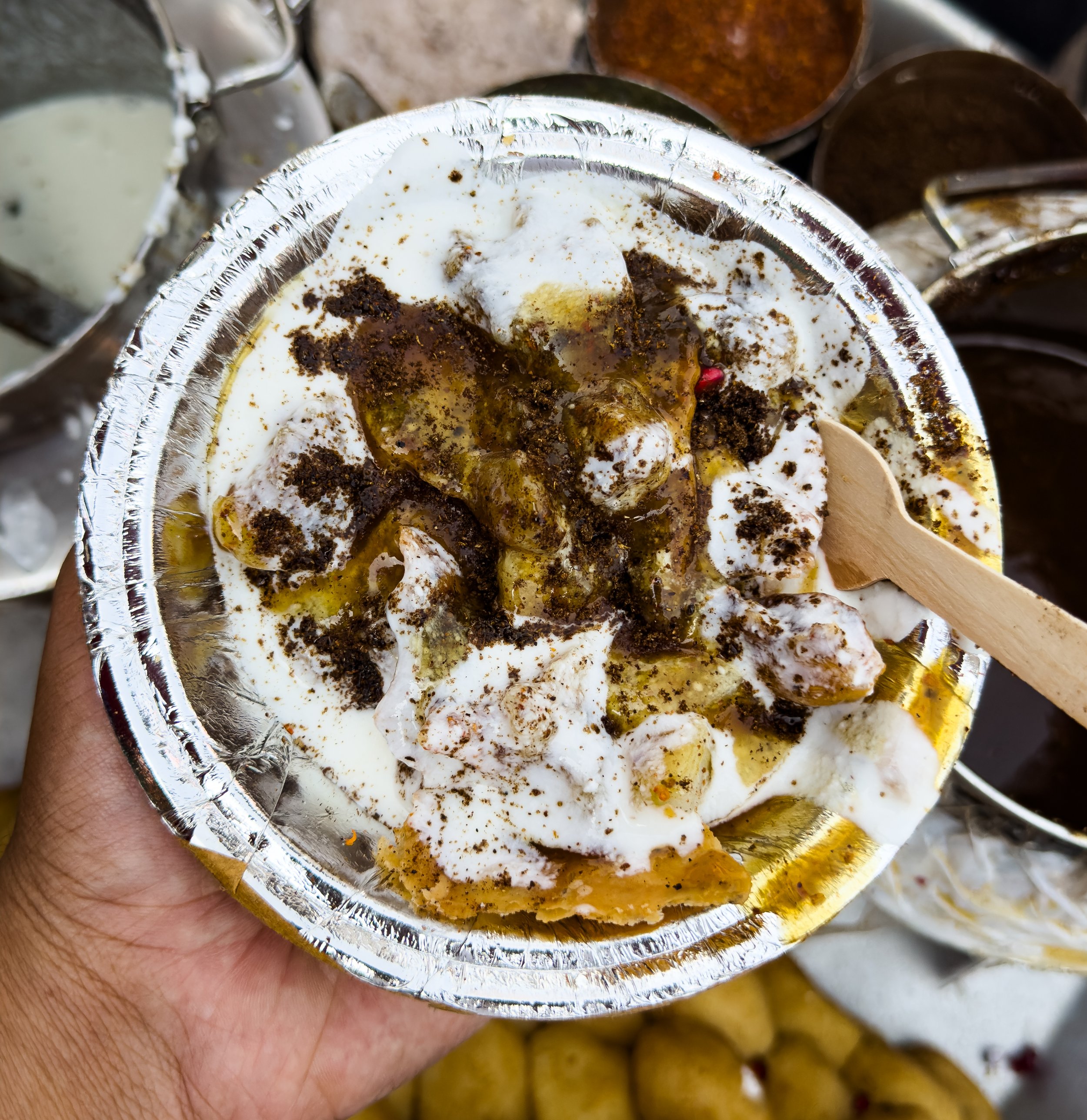 A hand holding a foil container filled with a Mexican dessert topped with whipped cream, chocolate sprinkles, and caramel sauce.