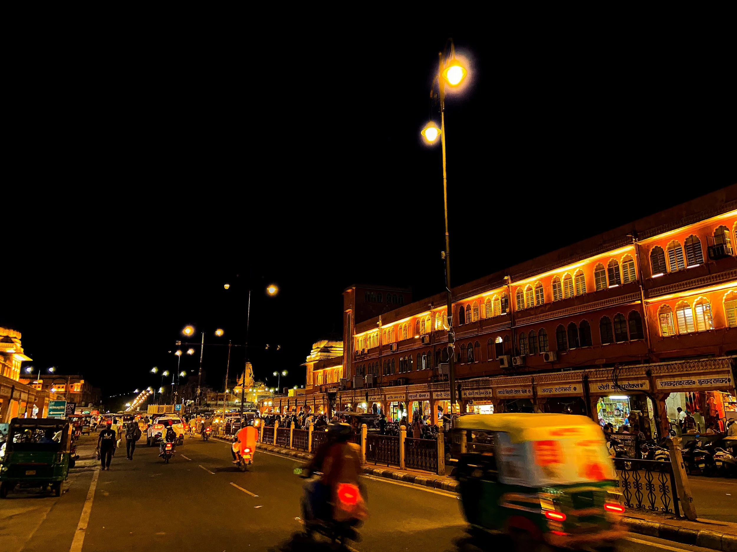 Nighttime street scene in India with illuminated historic buildings, streetlights, and moving vehicles including motorcycles and auto rickshaw.
