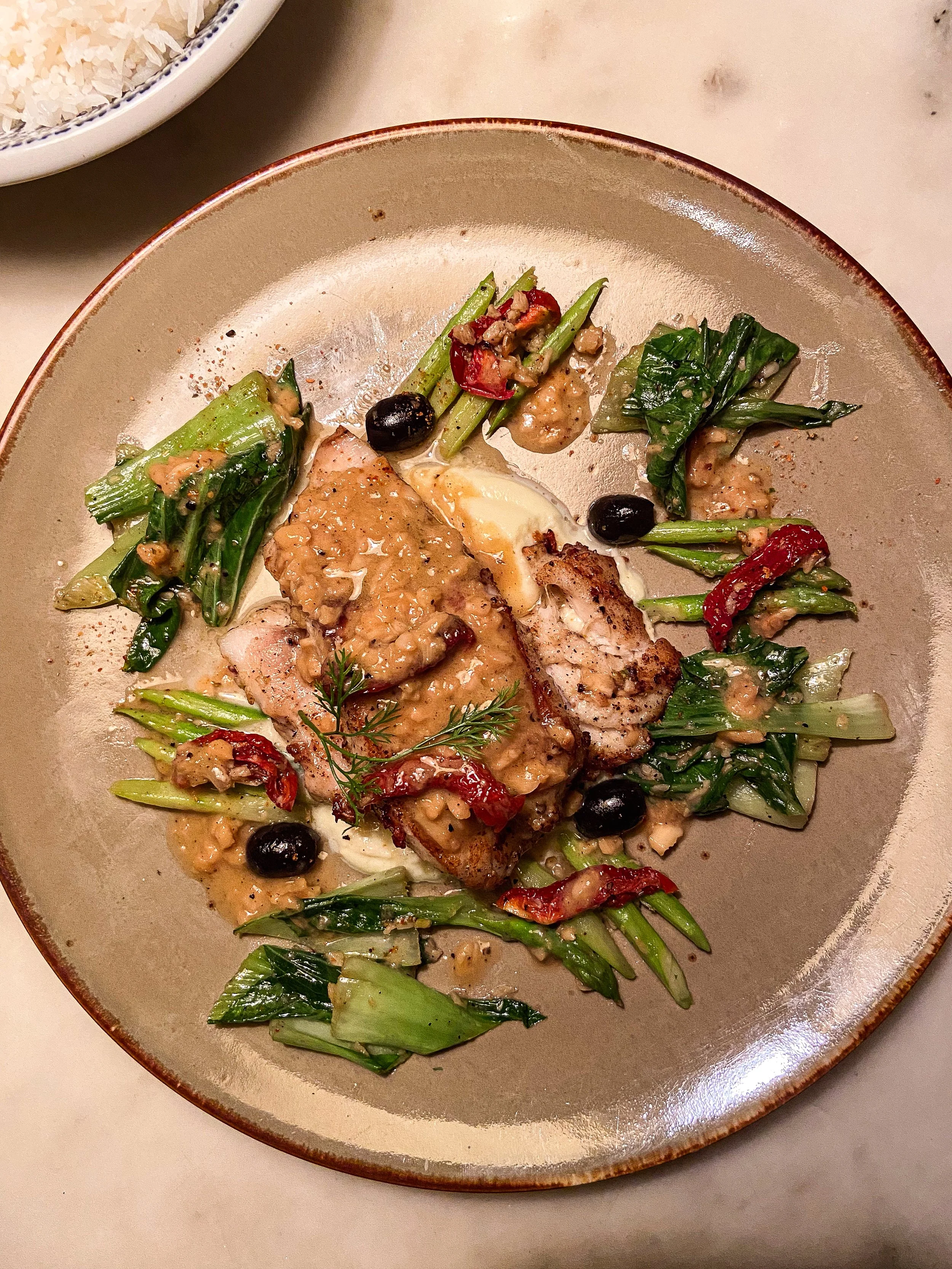 Plate of grilled chicken topped with sauce, served with sautéed greens, red peppers, black olives, and chopped nuts.
