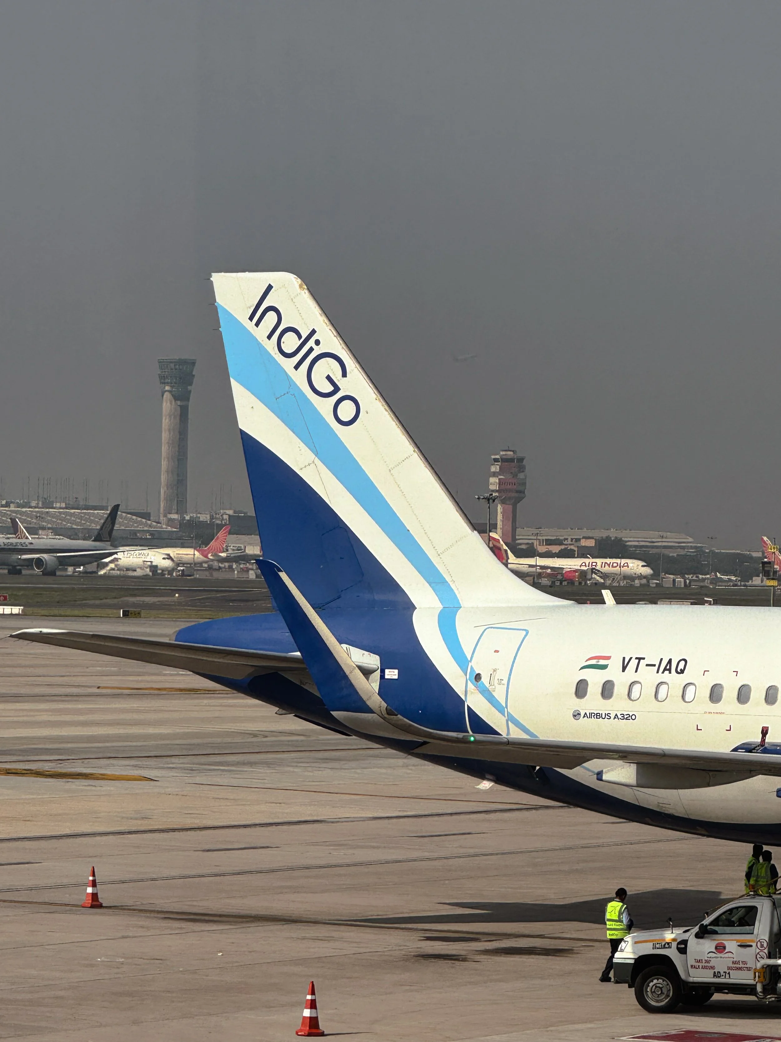 Close-up of the tail section of an IndiGo airplane on the tarmac, with other planes and airport structures visible in the background.