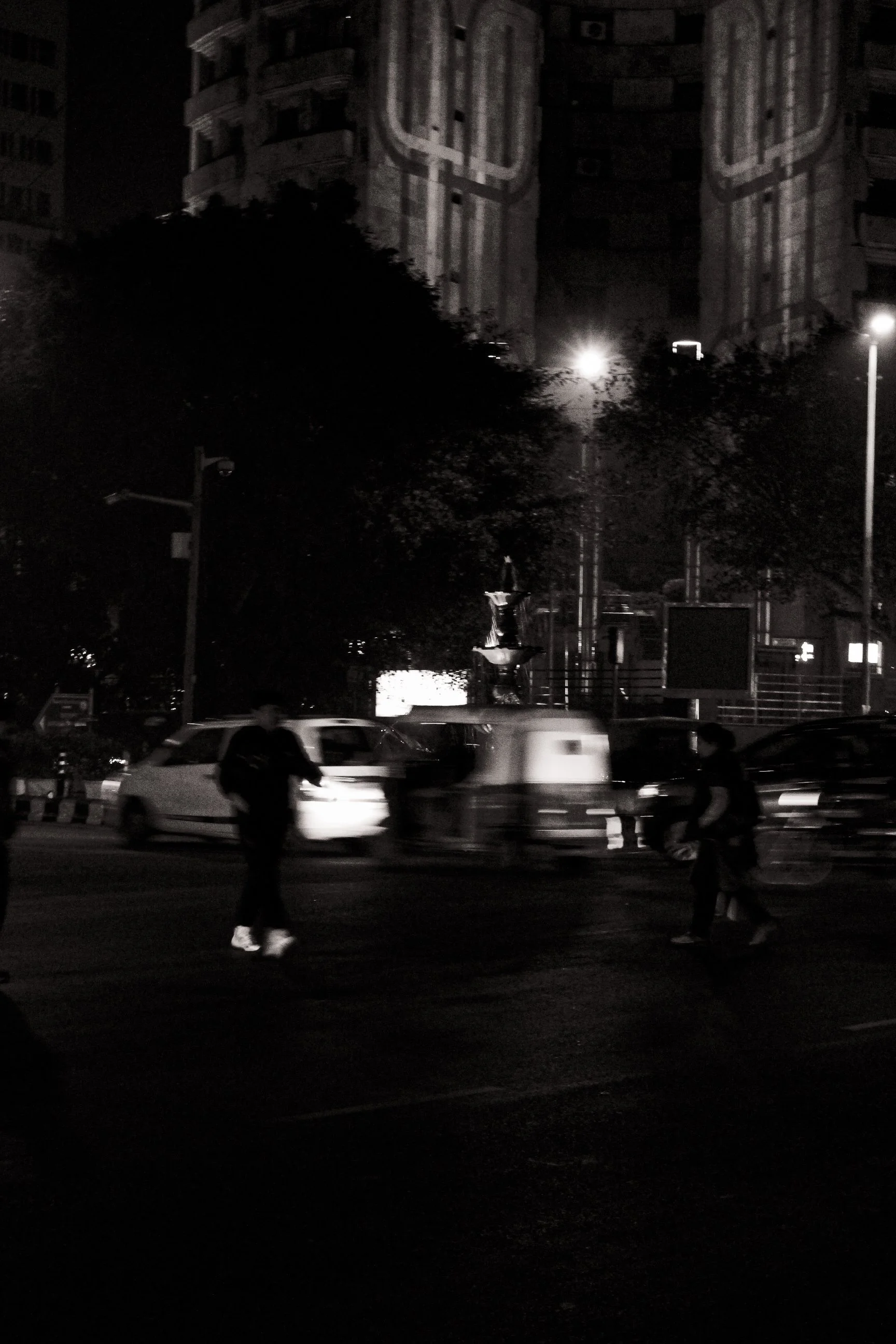 Nighttime city street scene with pedestrians crossing, cars, streetlights, and a large building with architectural details in the background.