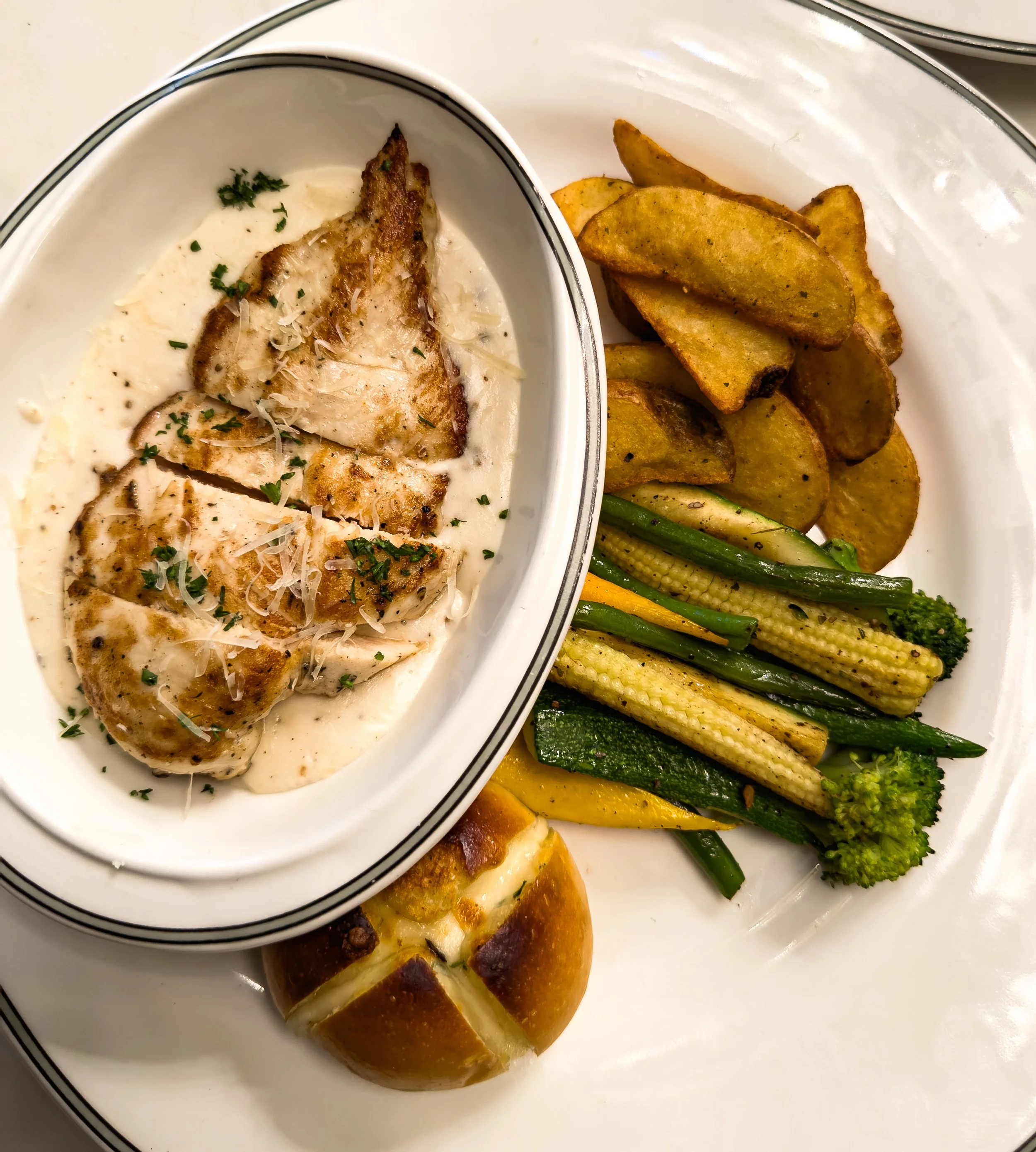A plate with grilled chicken in creamy white sauce, roasted potato wedges, steamed mixed vegetables including baby corn, green beans, zucchini, broccoli, and a small bread roll.