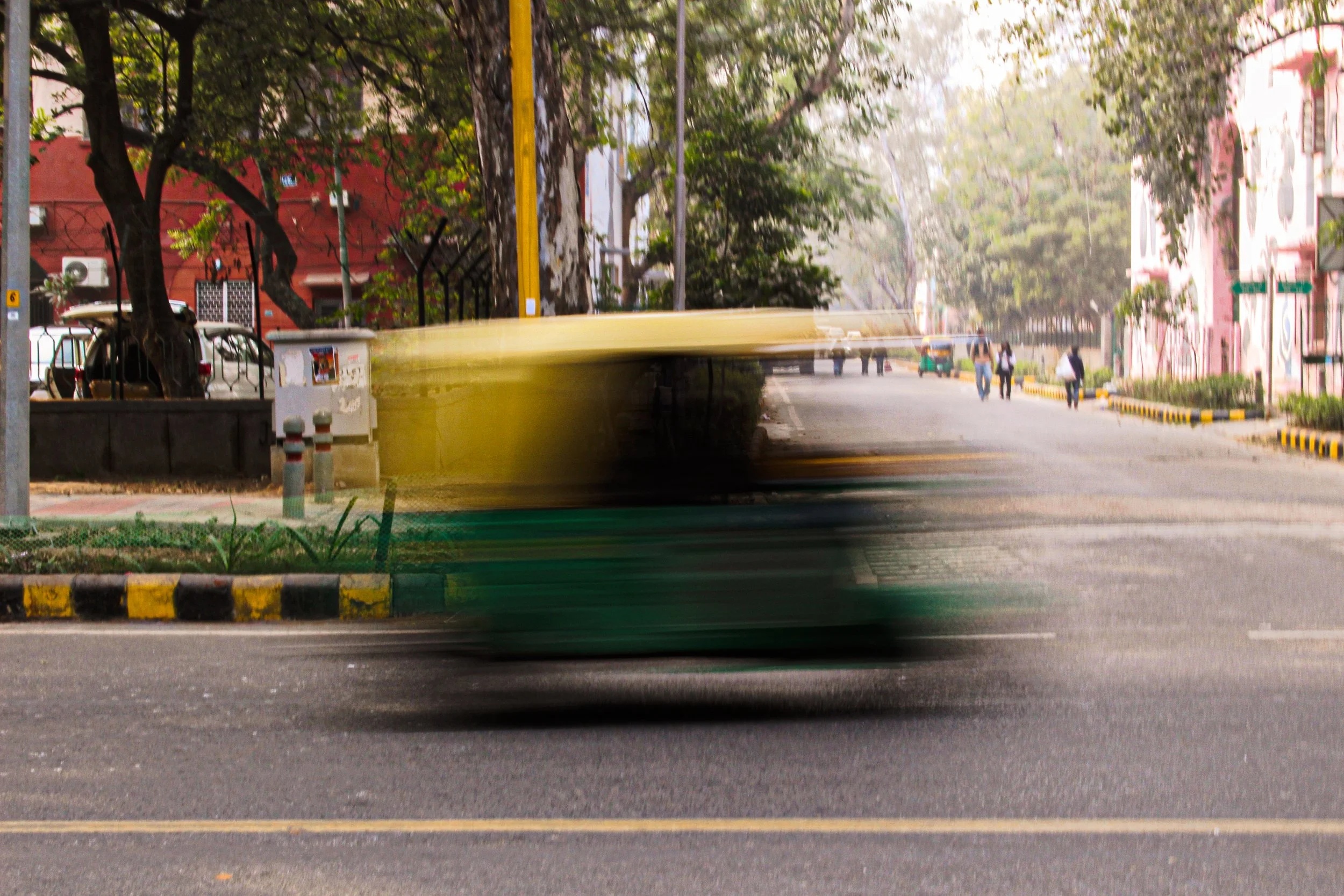 A blurred yellow and green vehicle moving quickly on a street in a city with trees, buildings, and pedestrians in the background.