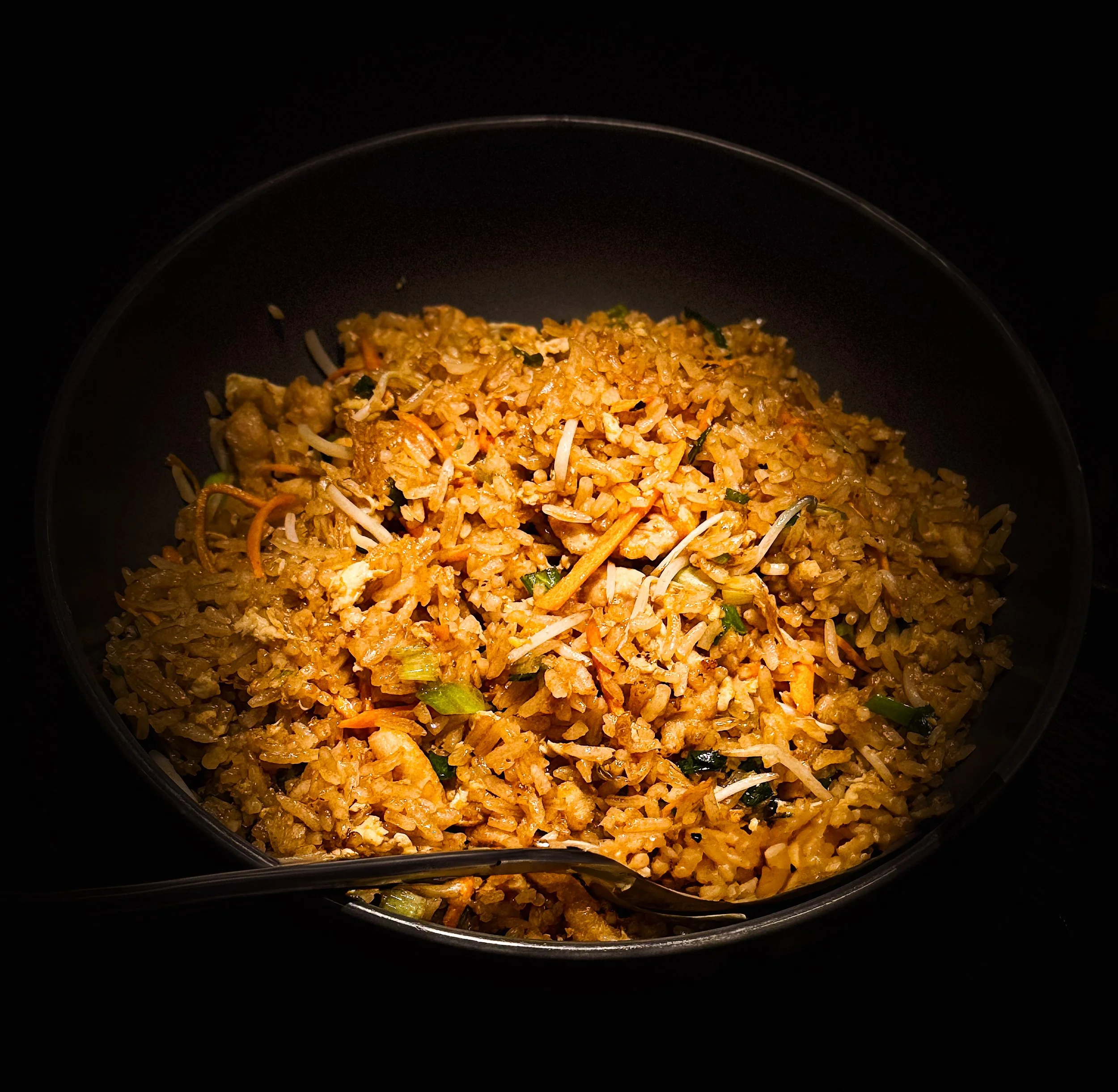 A bowl of fried rice with vegetables and scrambled eggs, served in a black bowl with a spoon.