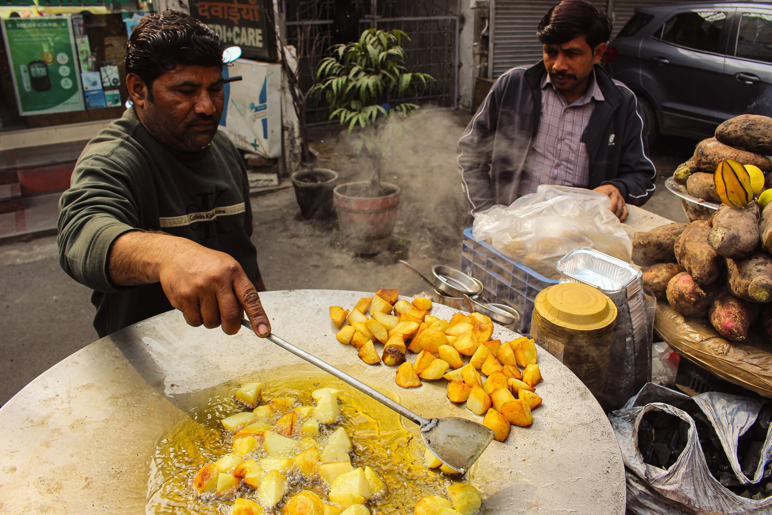 Two men cooking potatoes on a street food cart. One man is frying potatoes in oil, while the other man watches. There are sweet potatoes or yams stacked on a plate nearby, and various items like a plastic container and a cardboard box are on the cart