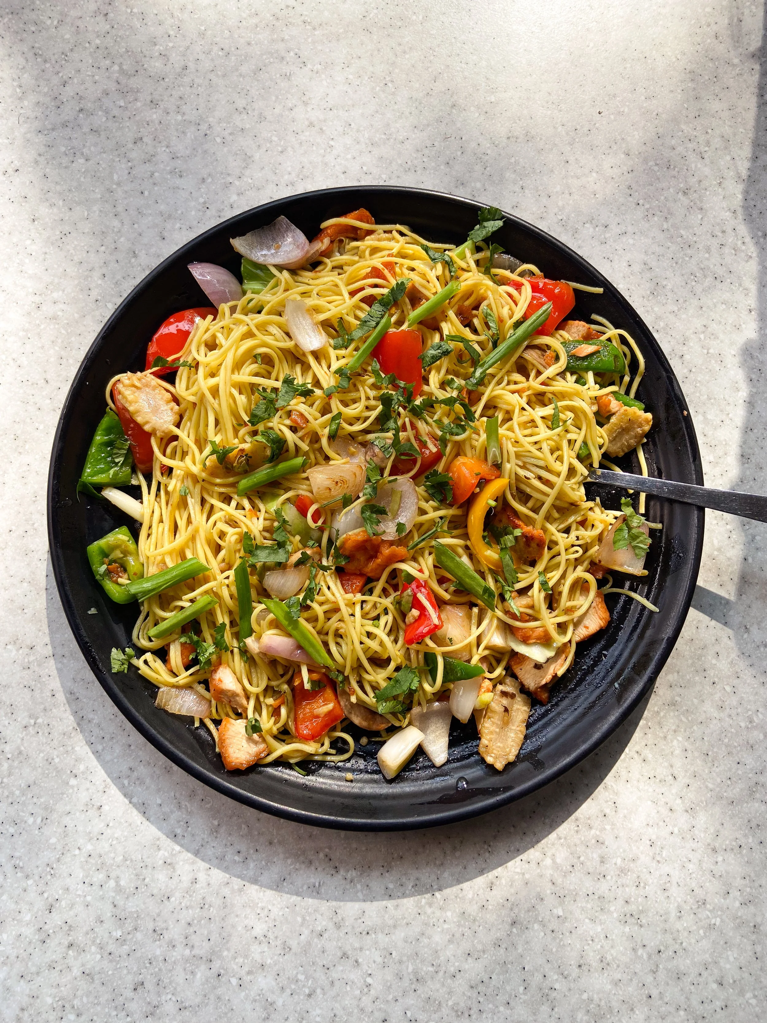 A black plate of stir-fried noodles with vegetables including bell peppers, onions, and green onions, garnished with chopped herbs.