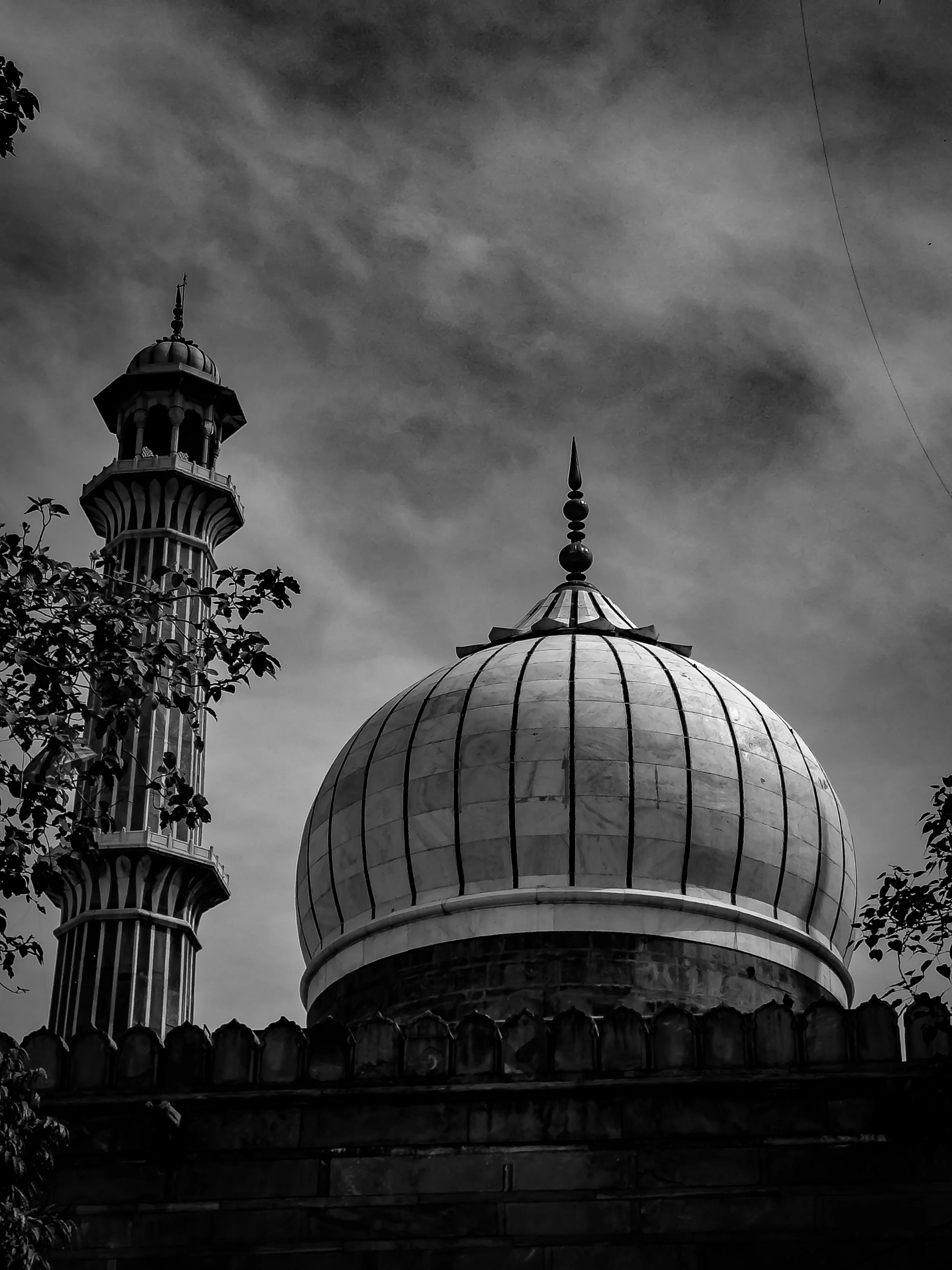 Black and white photo of a mosque with a large onion-shaped dome and a tall minaret, with trees and cloudy sky in the background.