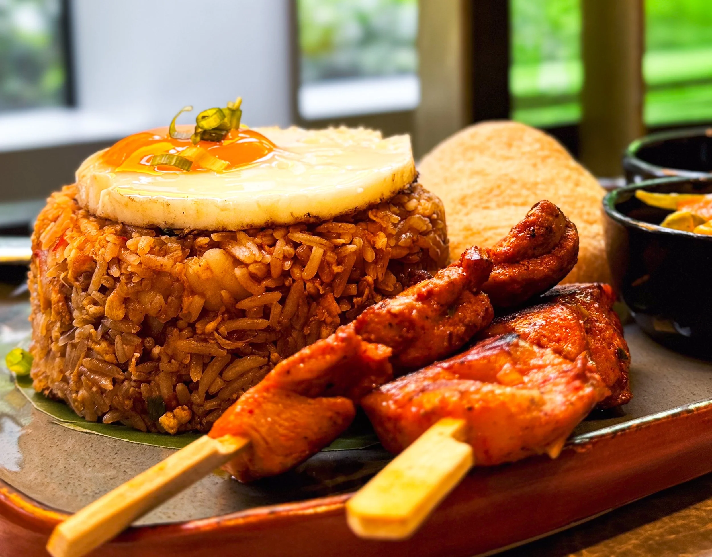 Plate of Jamaican jerk chicken, fried rice with a fried egg on top, plantain chips, and side dishes in black bowls on a wooden table.
