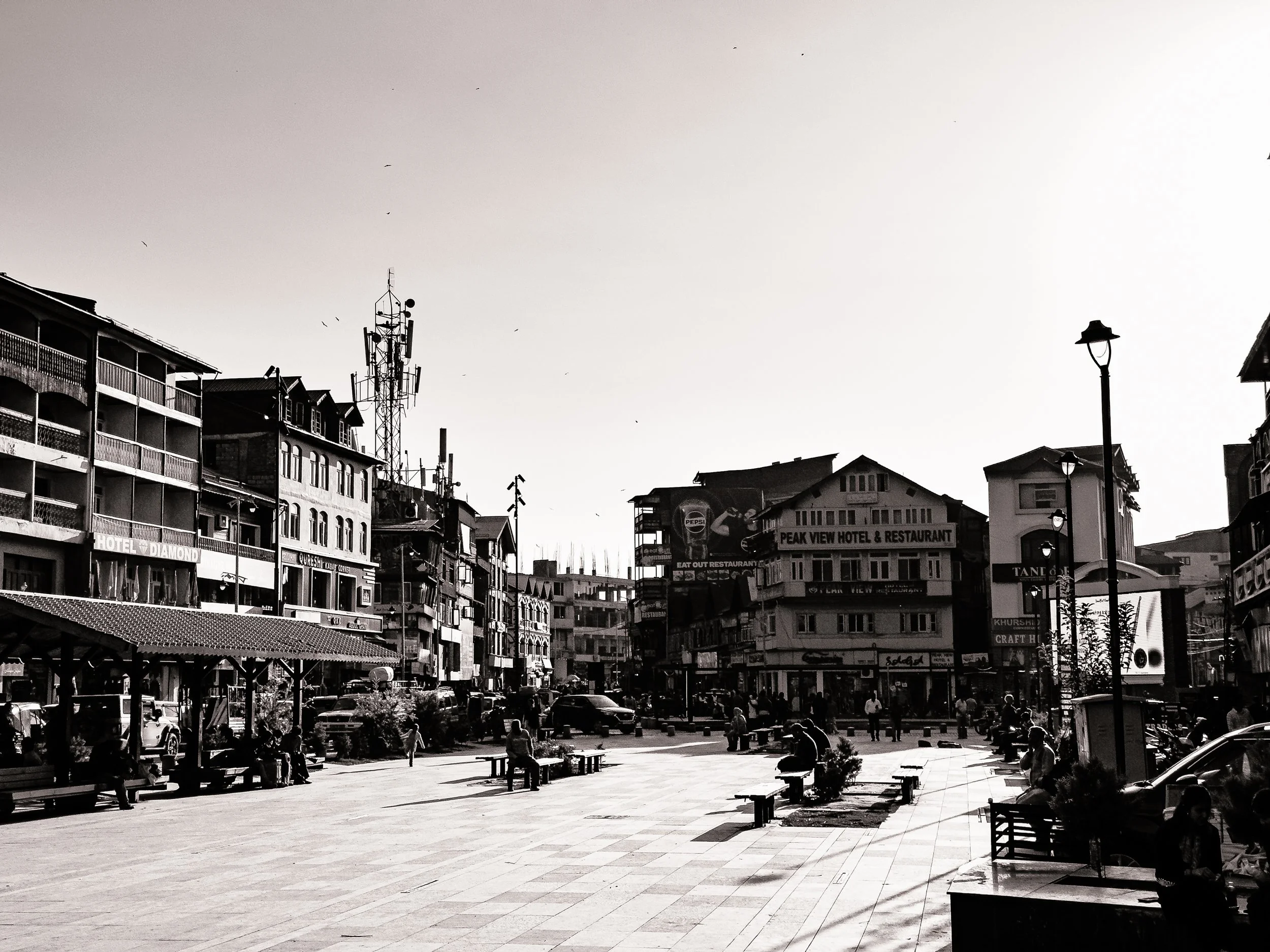 Black and white photo of a busy urban square with benches, people walking, and multi-story buildings including a hotel, restaurant, and shops. Streets filled with parked cars and a communication tower in the background.