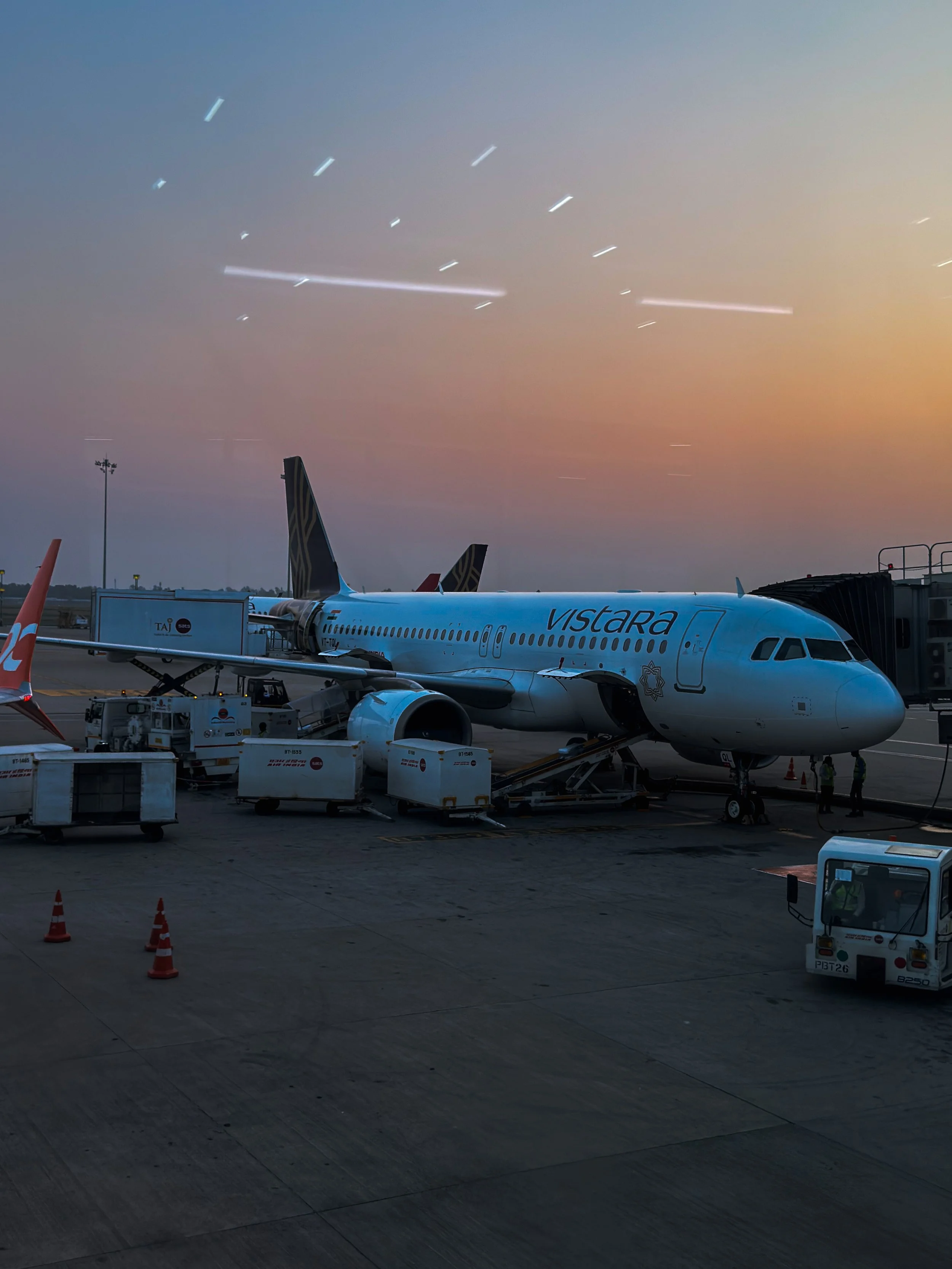 An airplane with 'Vistara' written on its side parked at the gate during sunset, with streaks of light in the sky caused by moving aircraft lights.