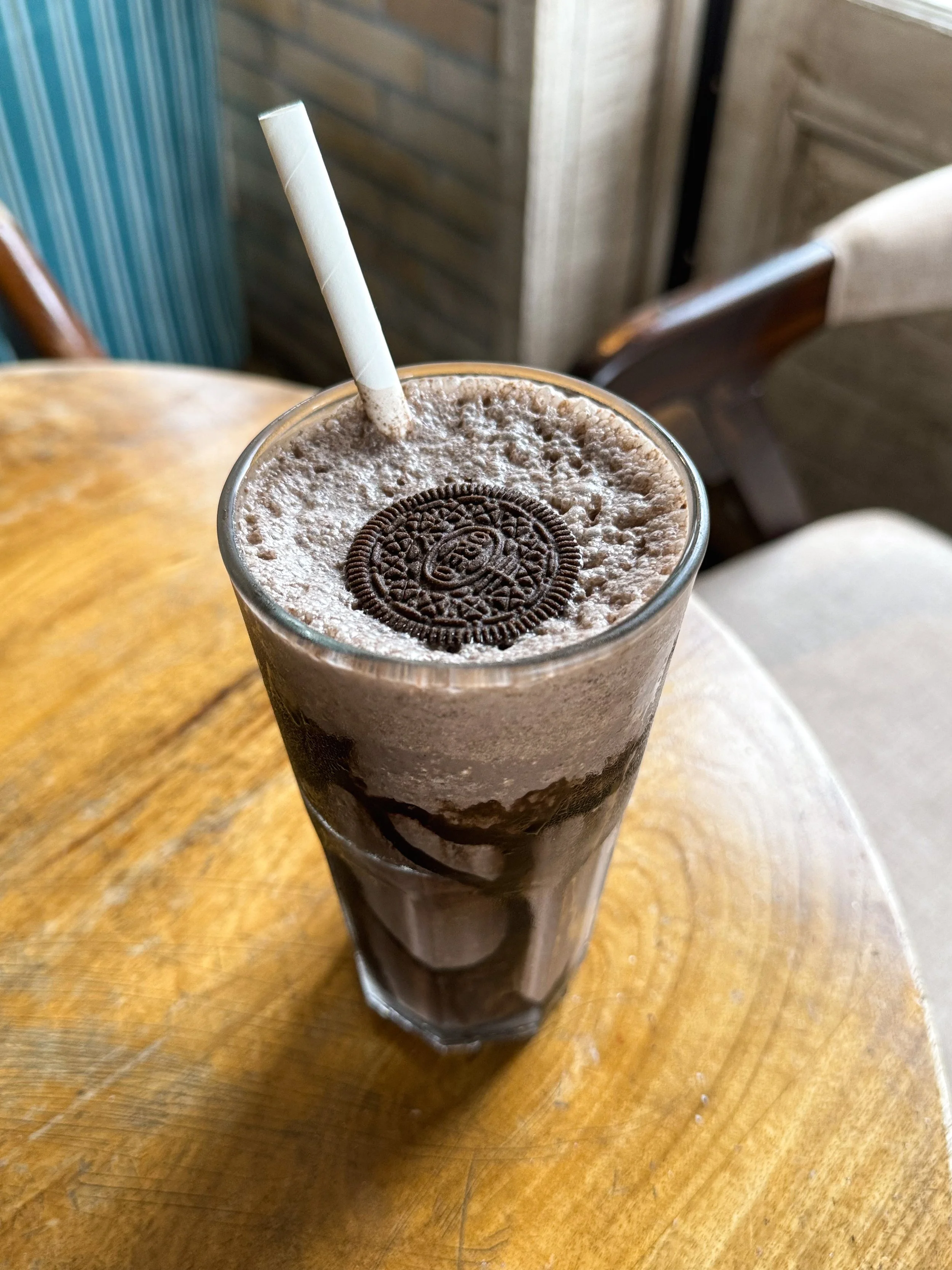A tall glass of chocolate milkshake topped with whipped cream and an Oreo cookie, with a striped paper straw, on a wooden table.