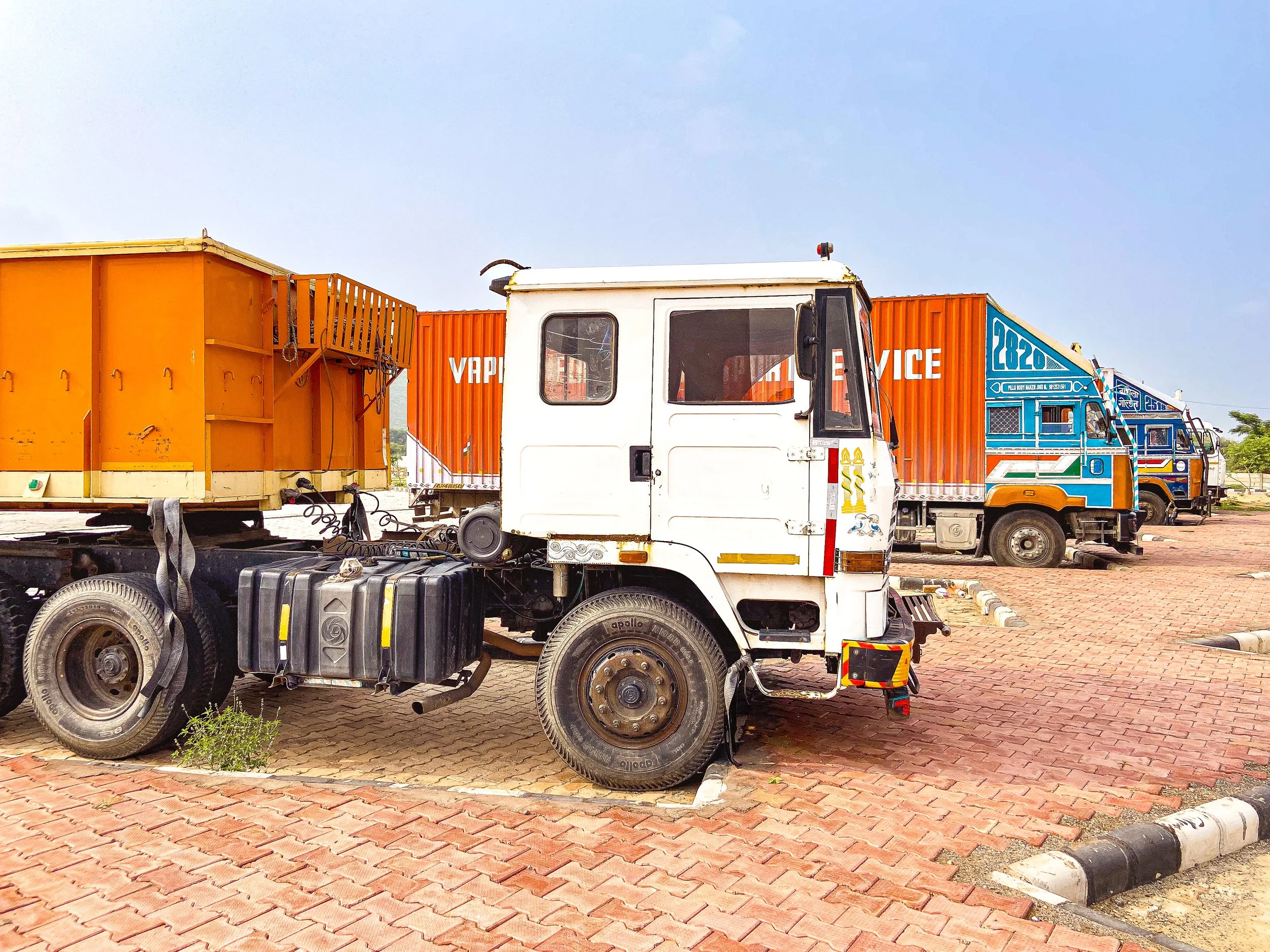 A white truck parked on a brick-paved area, with colorful trucks and containers in the background.
